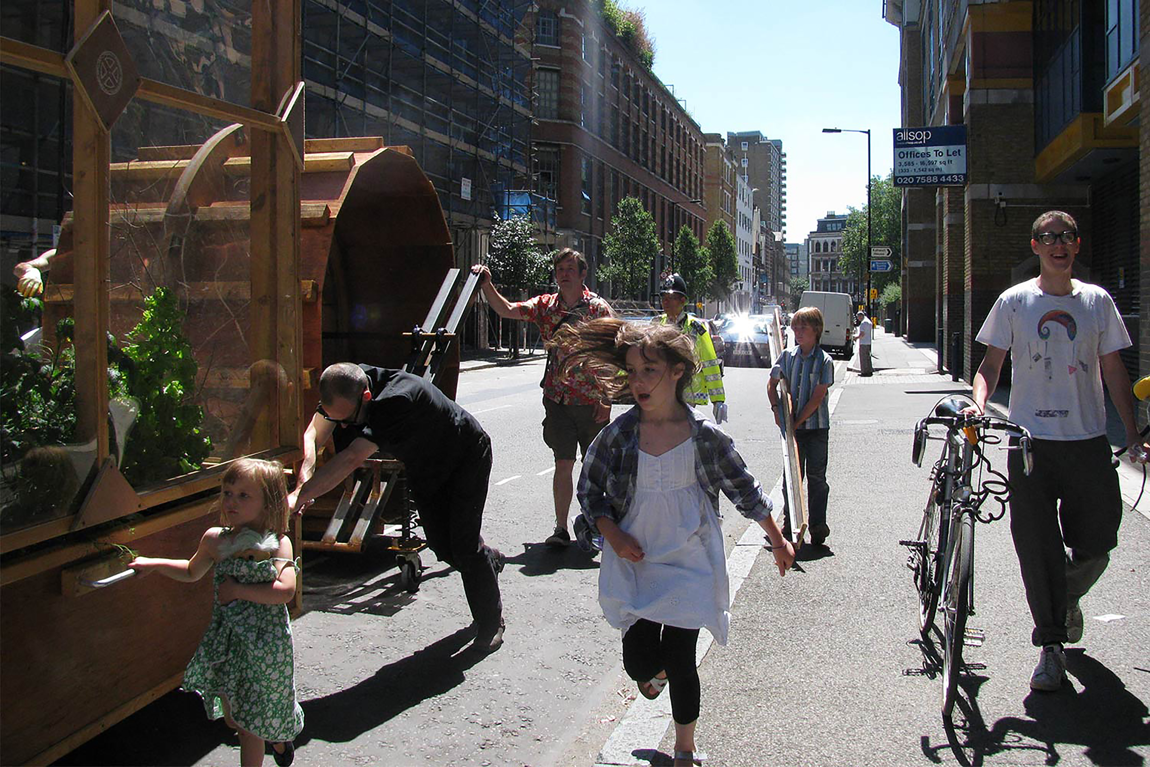 A photograph of two girls running alongside a wooden framed green house being pushed by a large wooden wheel.