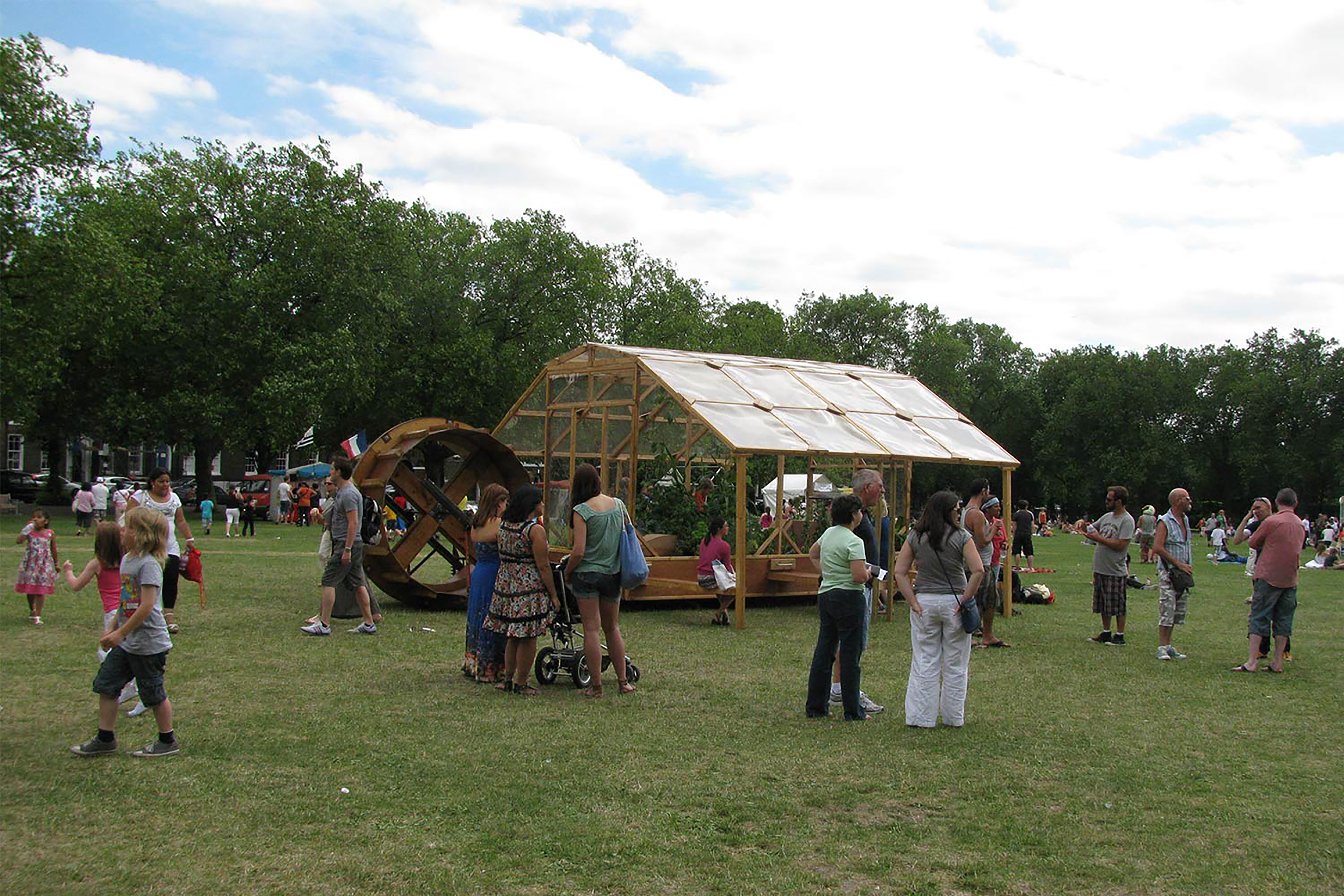 A photograph of a wooden framed green house in the middle of a field surrounded by crowds.