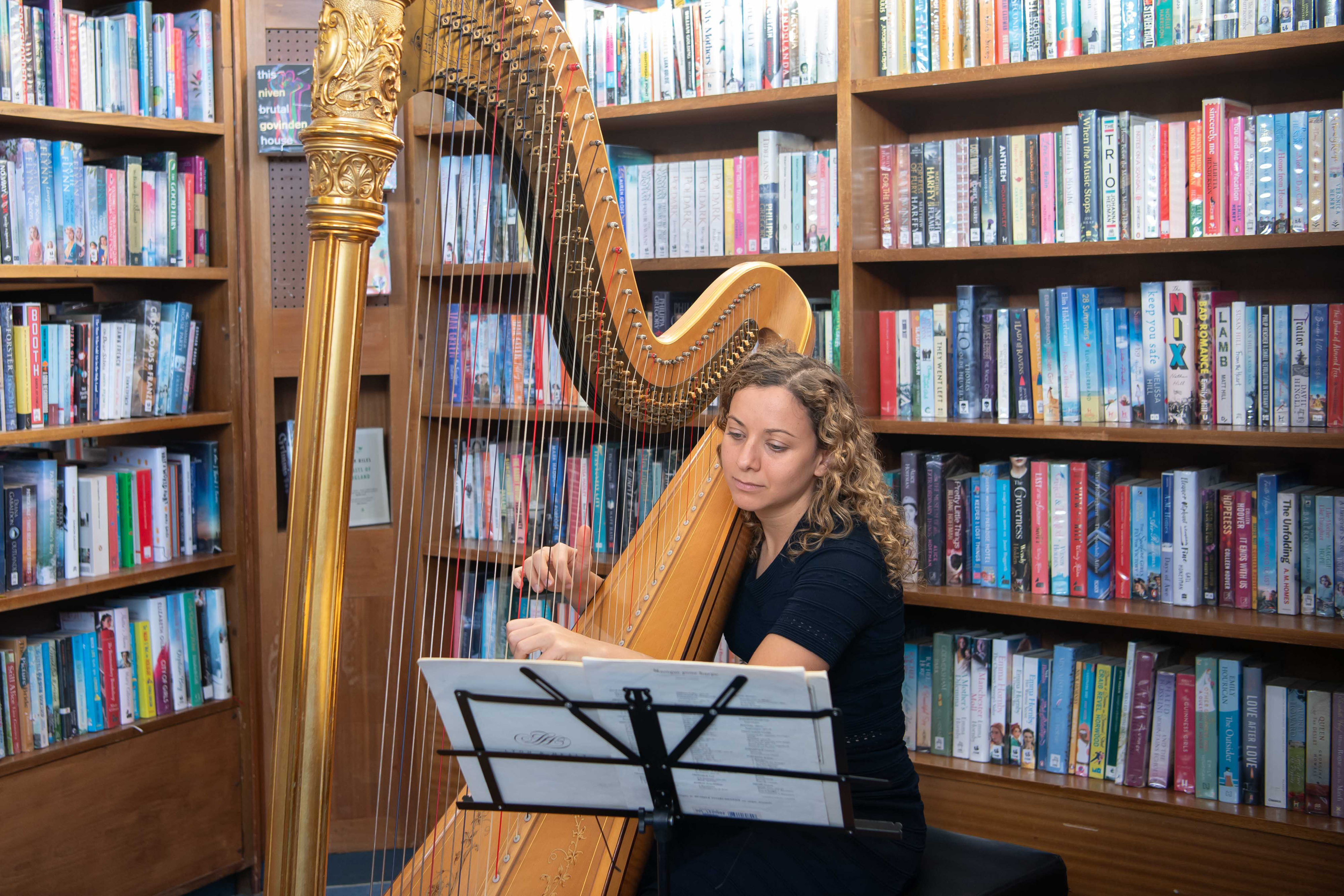 A woman playing a large harp amongst a wall of library books on shelves. She is sitting down next to a music stand with paper on it.
