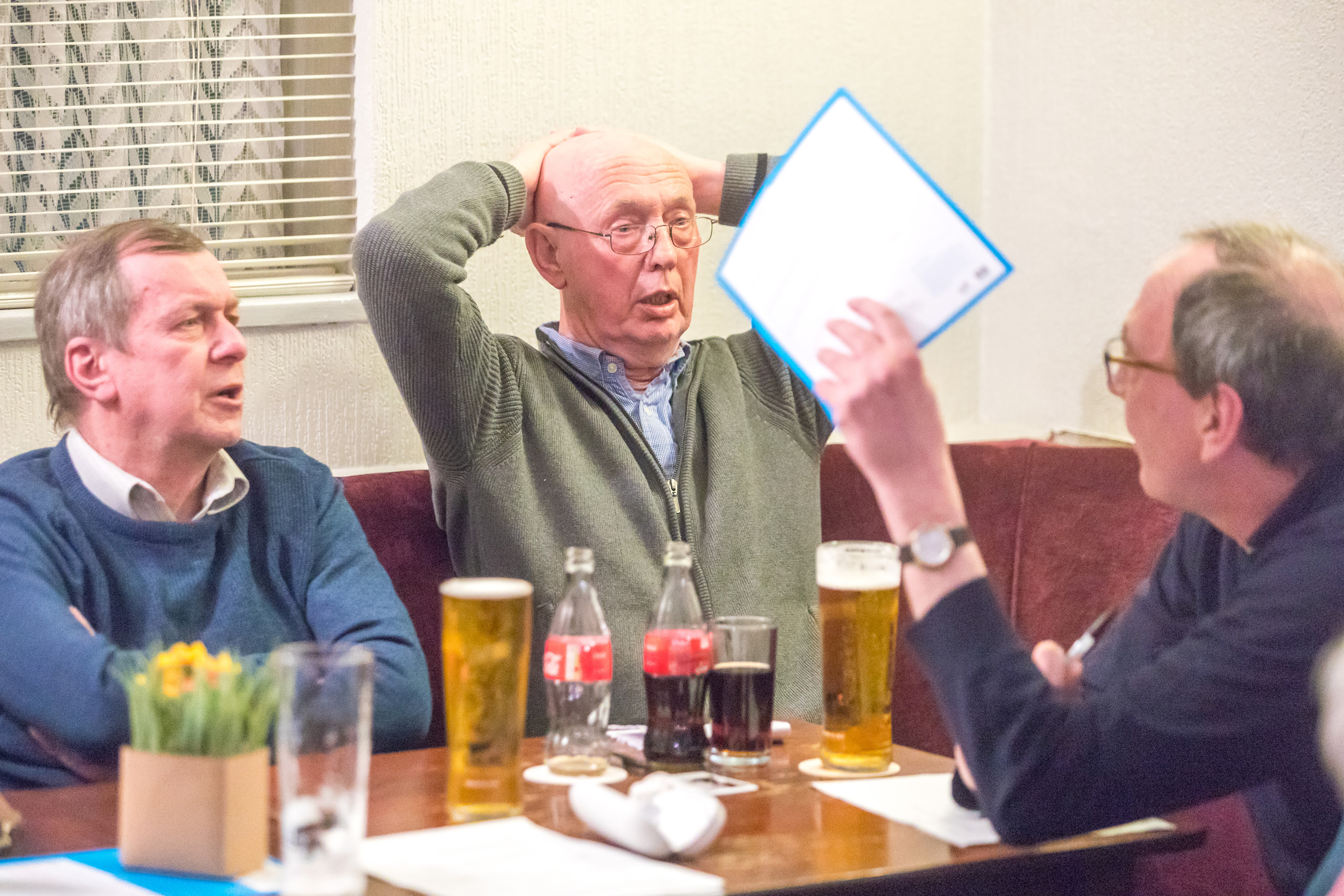 Three middle-aged white men talking in a pub with beers cluttering a table.