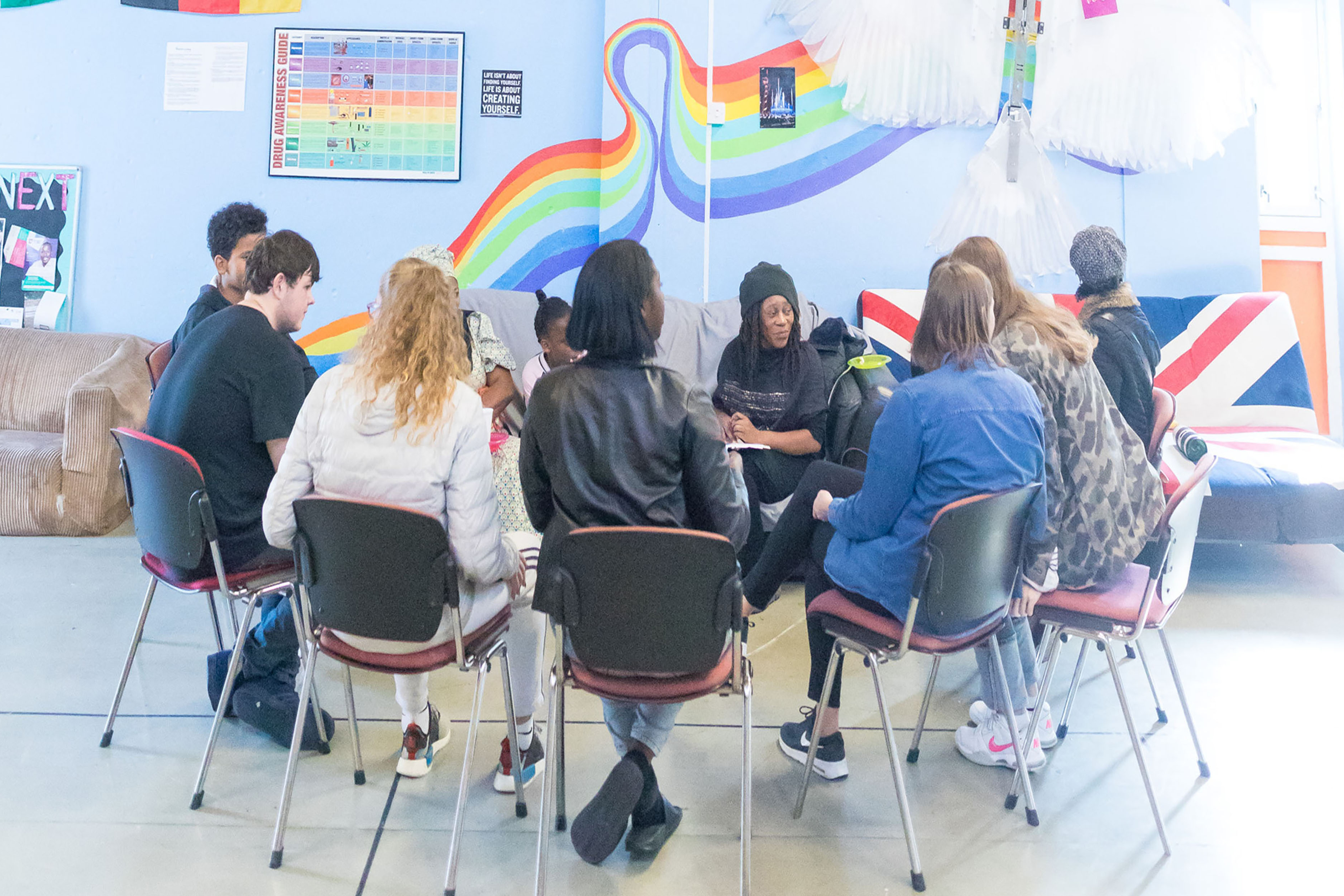 A photograph of a group of teenagers sitting in chairs forming a circle in front of a colourfully decorated wall.