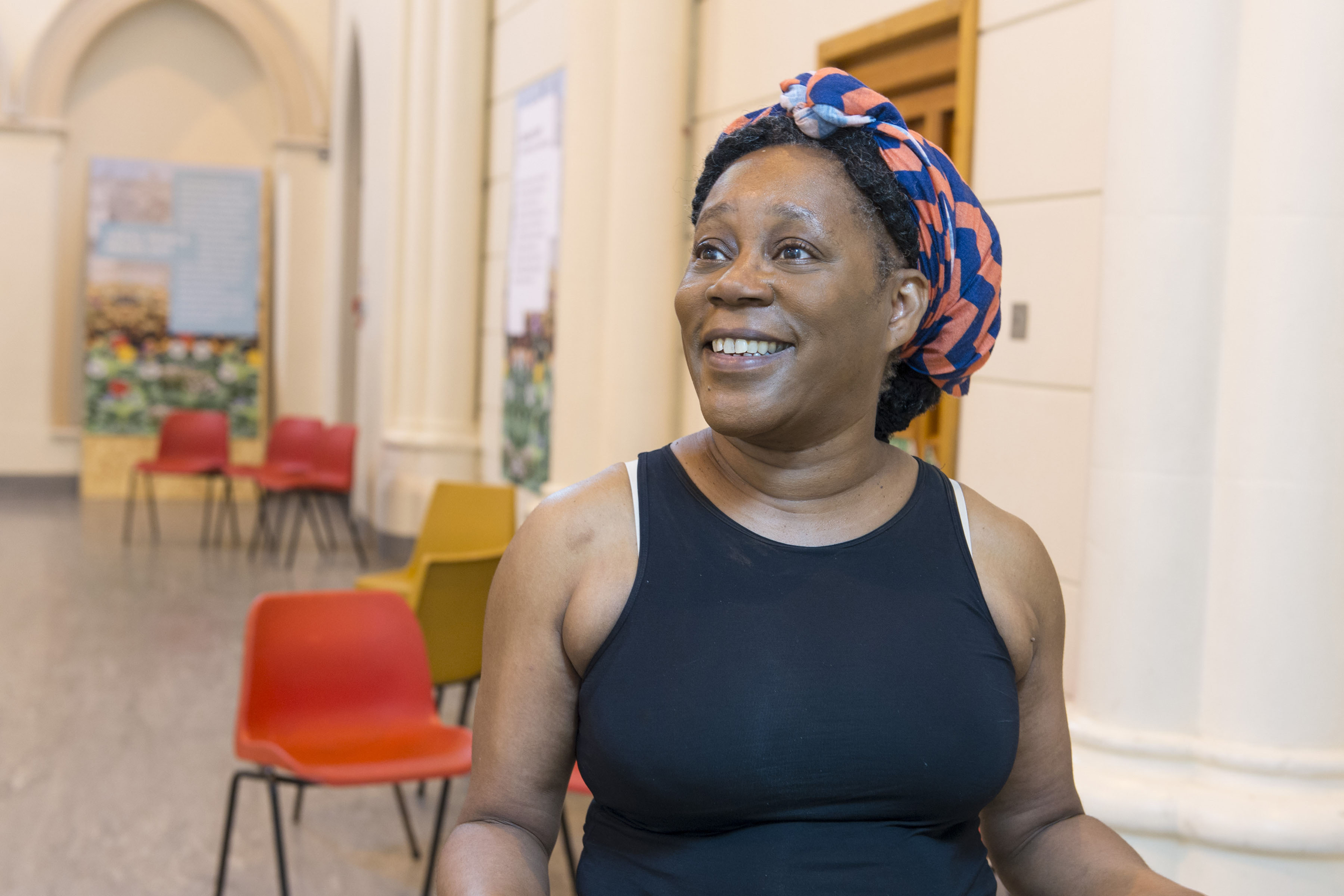 A photograph of a middle aged black woman smiling wearing a black tank top and a colourful hair wrap sitting on a chair in an open hall with chairs and artworks behind her.