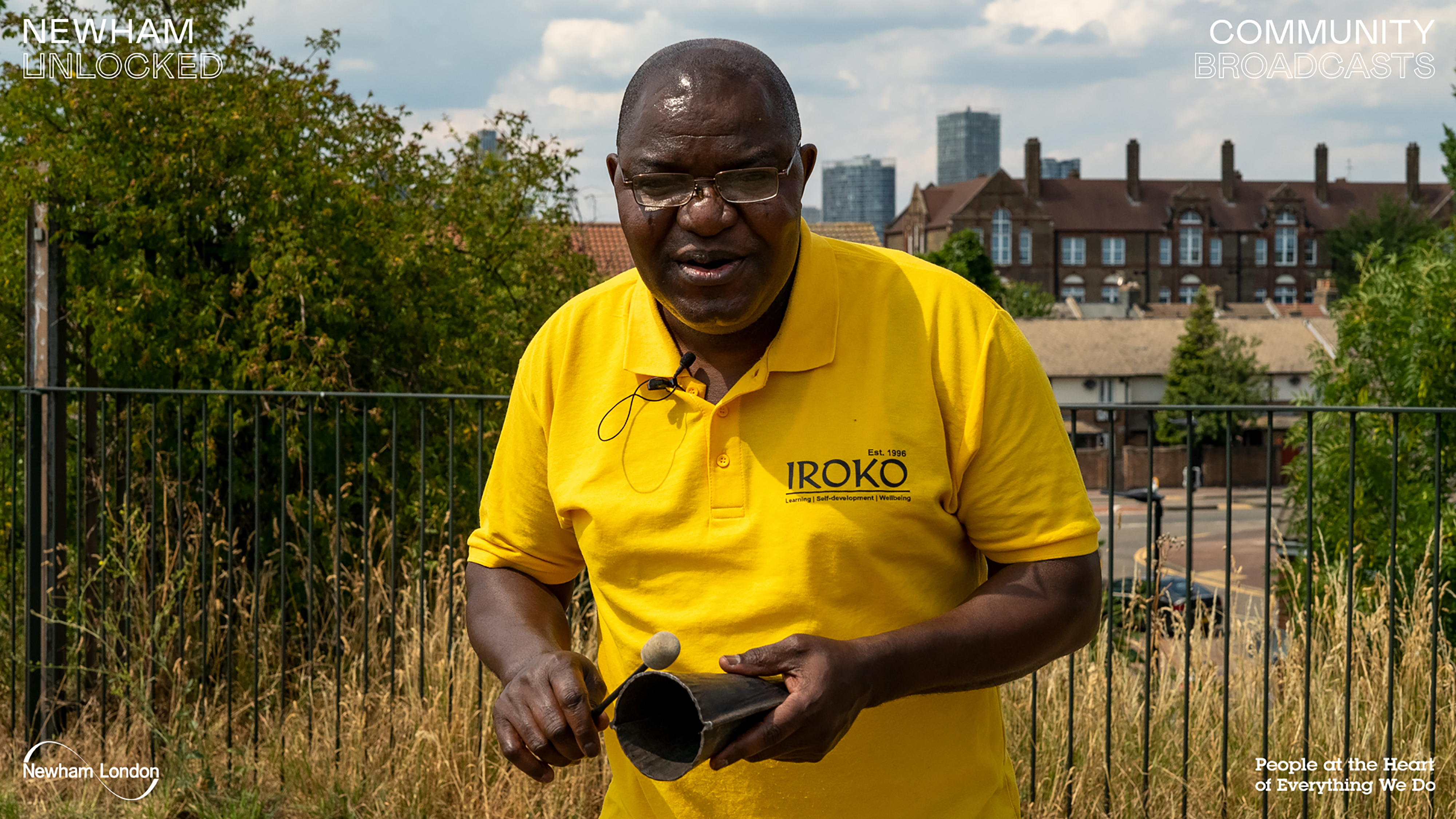 A photograph of a middle-aged black man wearing a yellow shirt playing a triangle in front a gated community.