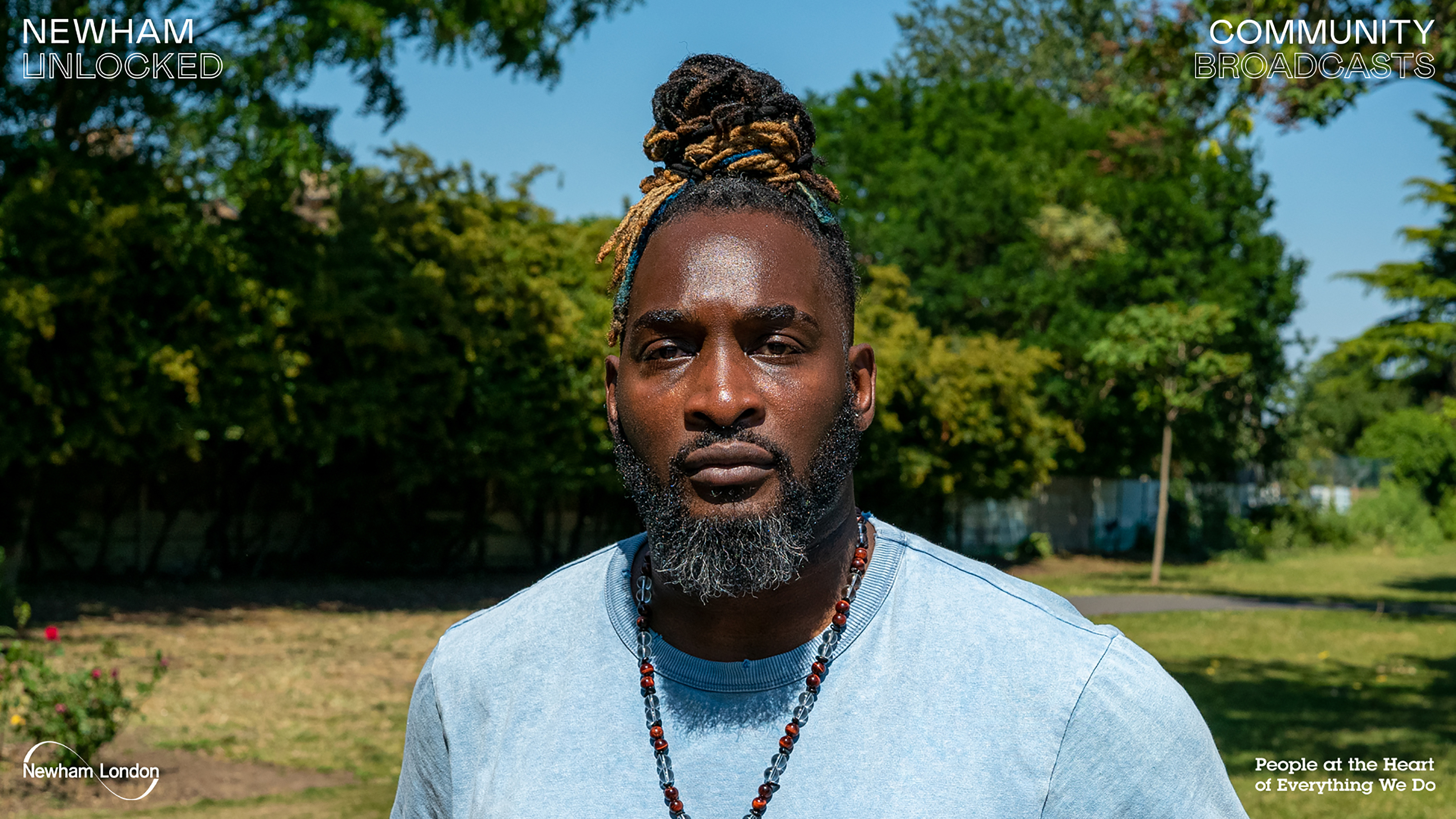 A photograph of a black man with dreads tied in a bun wearing a beaded necklace standing in front a park.