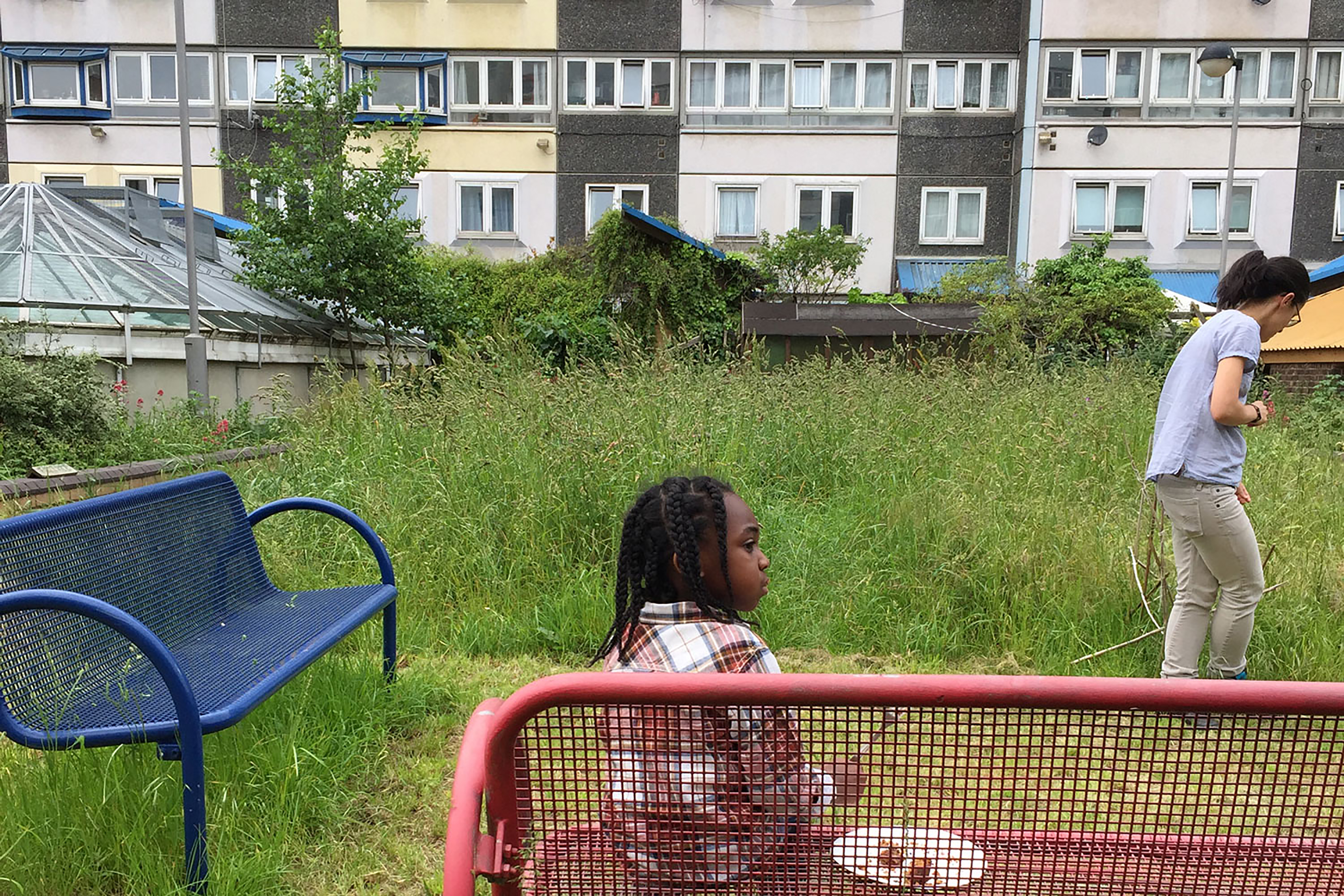 A young child sitting down on a red bench wearing a checked shirt, while a woman maintains the overgrown communal garden space in front of a row of large tower block homes.