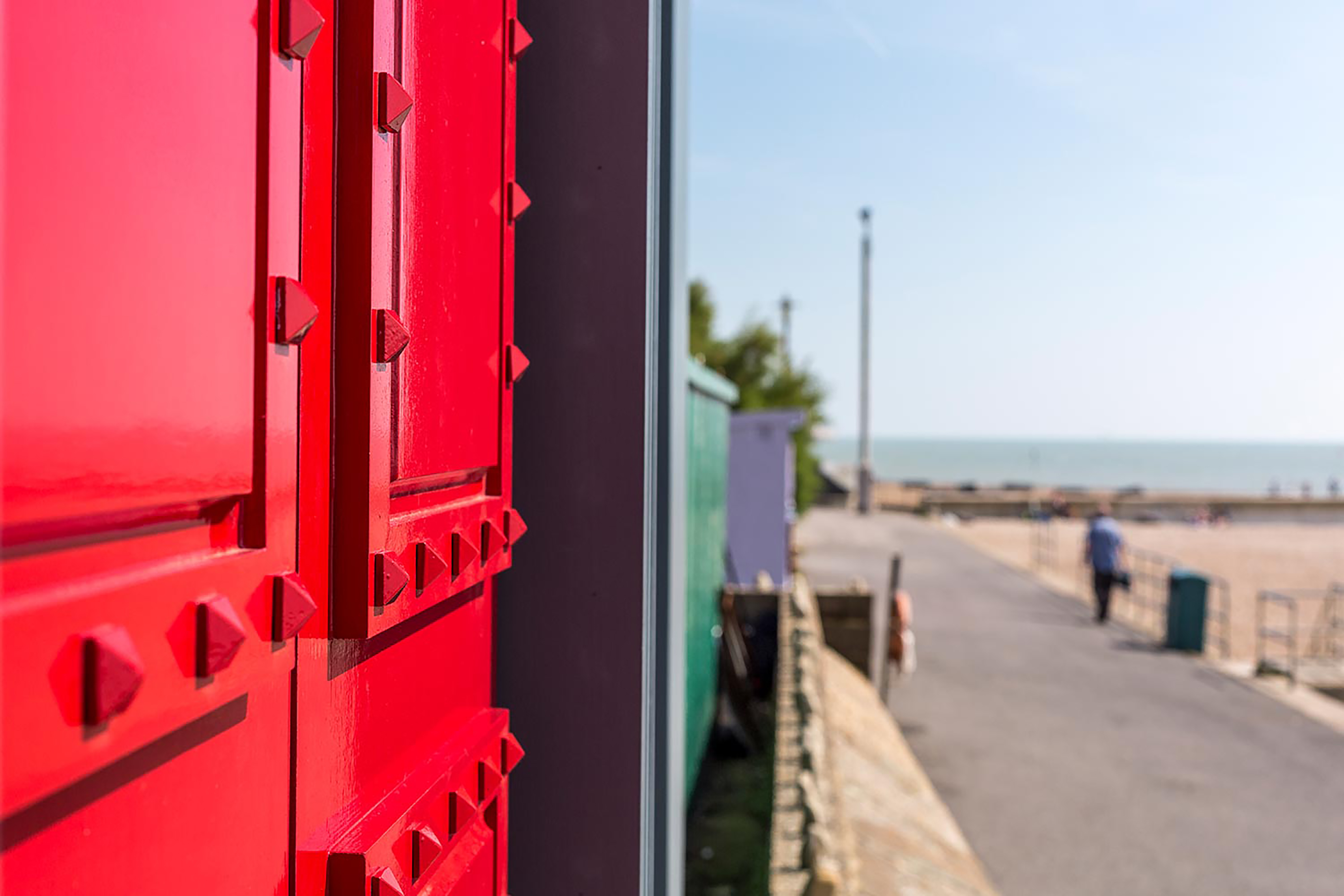 A close-up photograph of a detailed red beach hut door, side on with an out of focus beach walkway in the background.