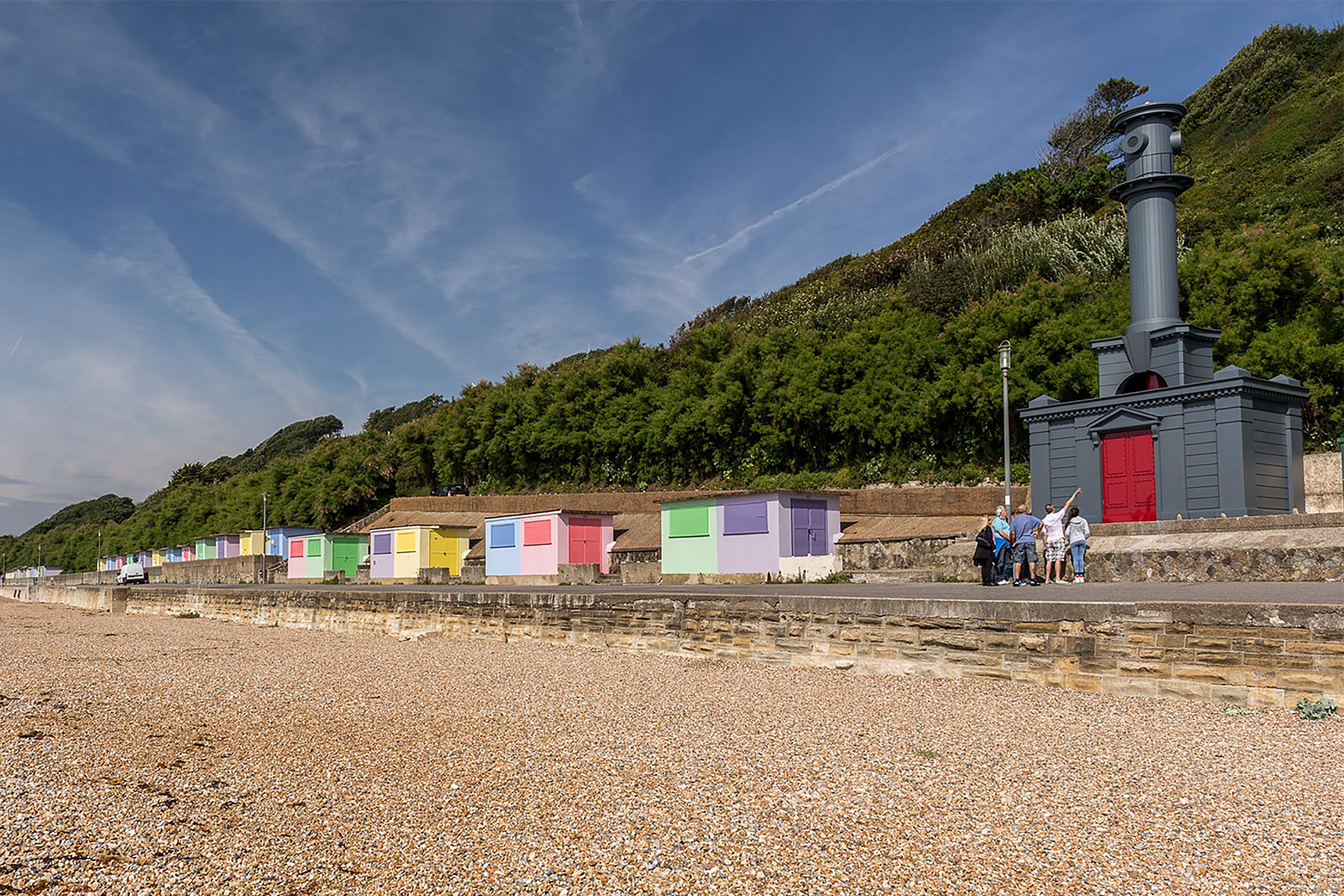 A photograph of a crowd gathering round a dark grey Hawksmoor beach hut along with a row of colourful beach huts spread across the coastline.