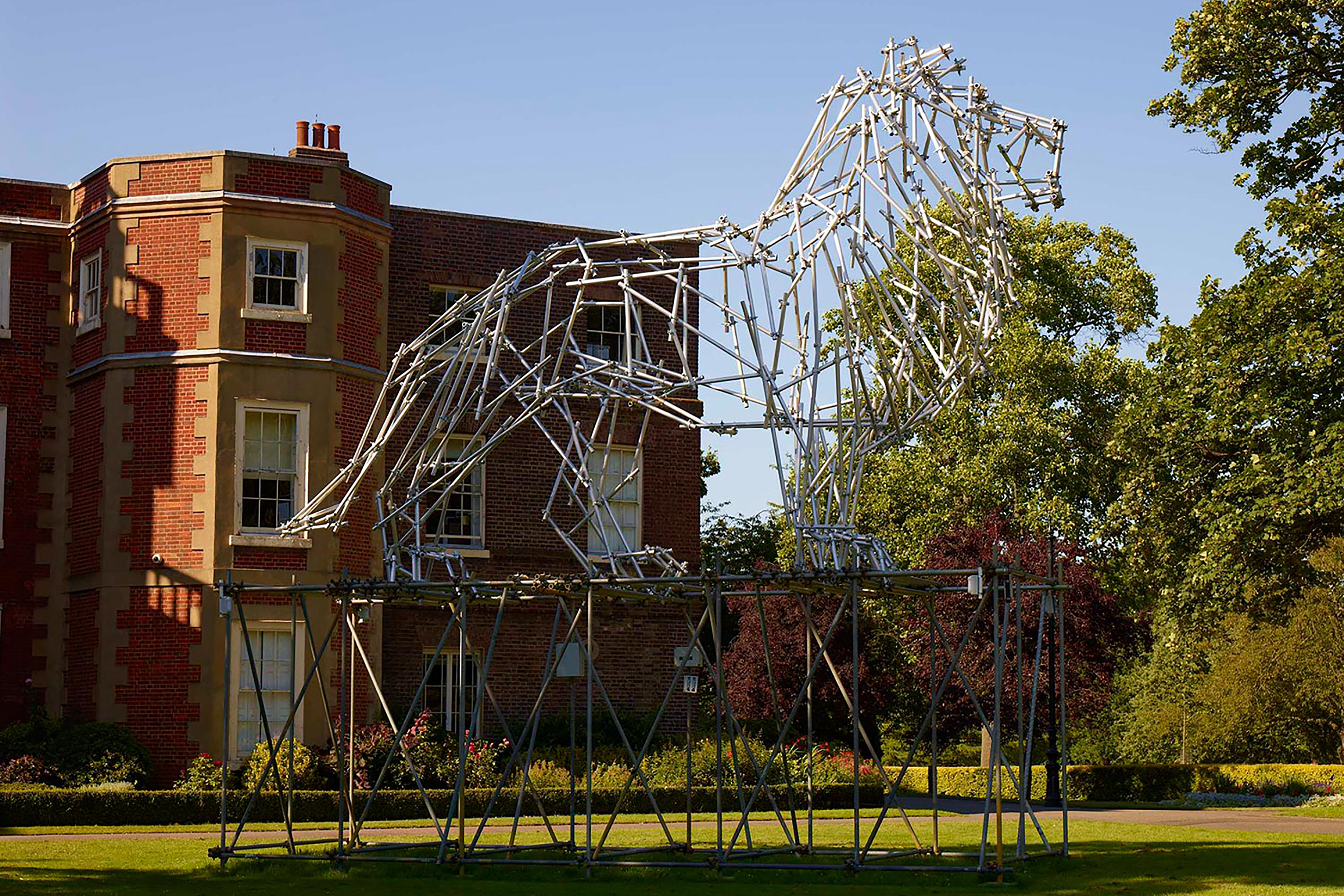 A large structure of a lion made up from small metal poles, on a large platform outside of a large red-brick building.