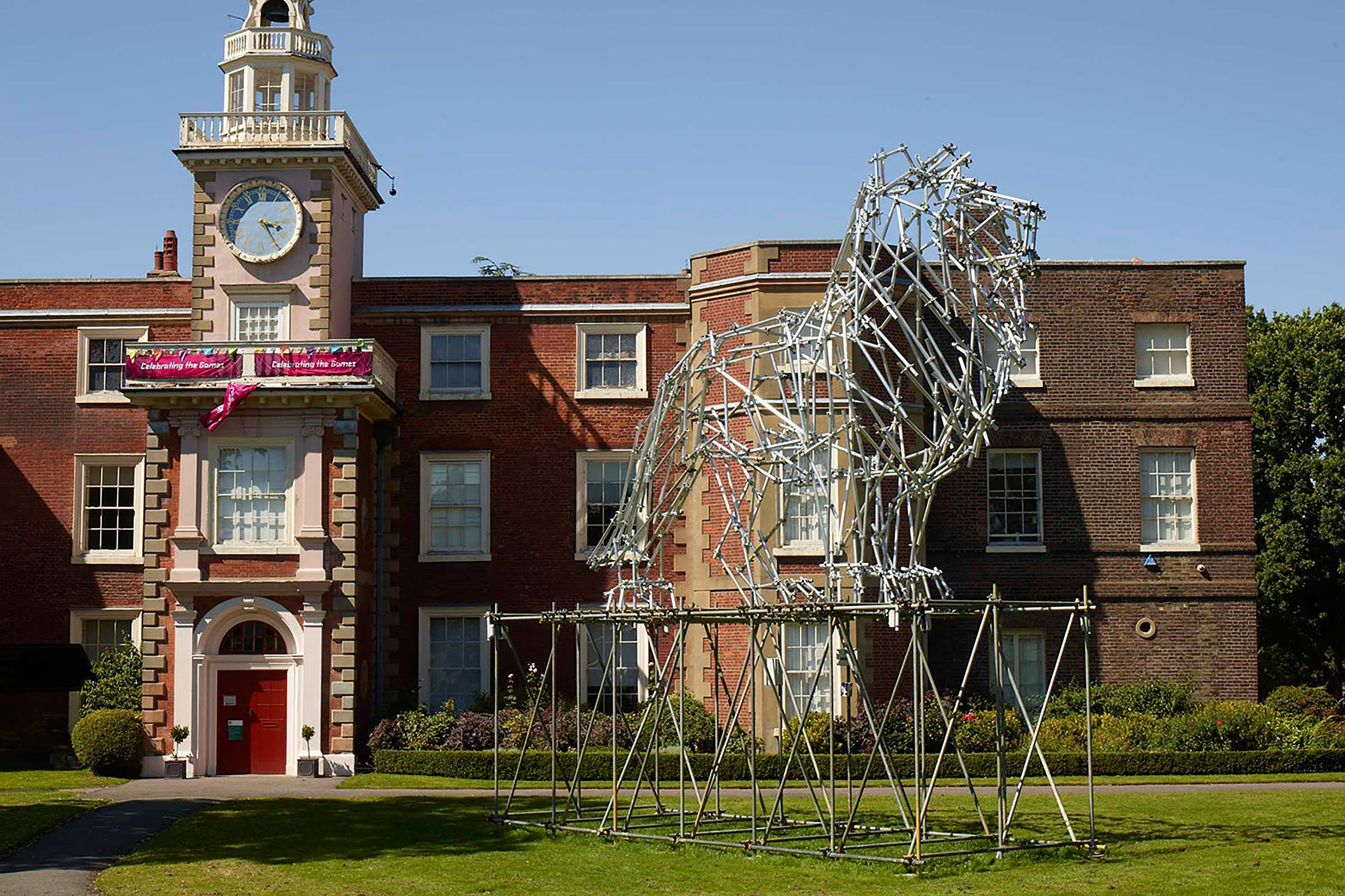 A large structure of a lion made up from small metal poles, on a large platform outside of a large red-brick building.