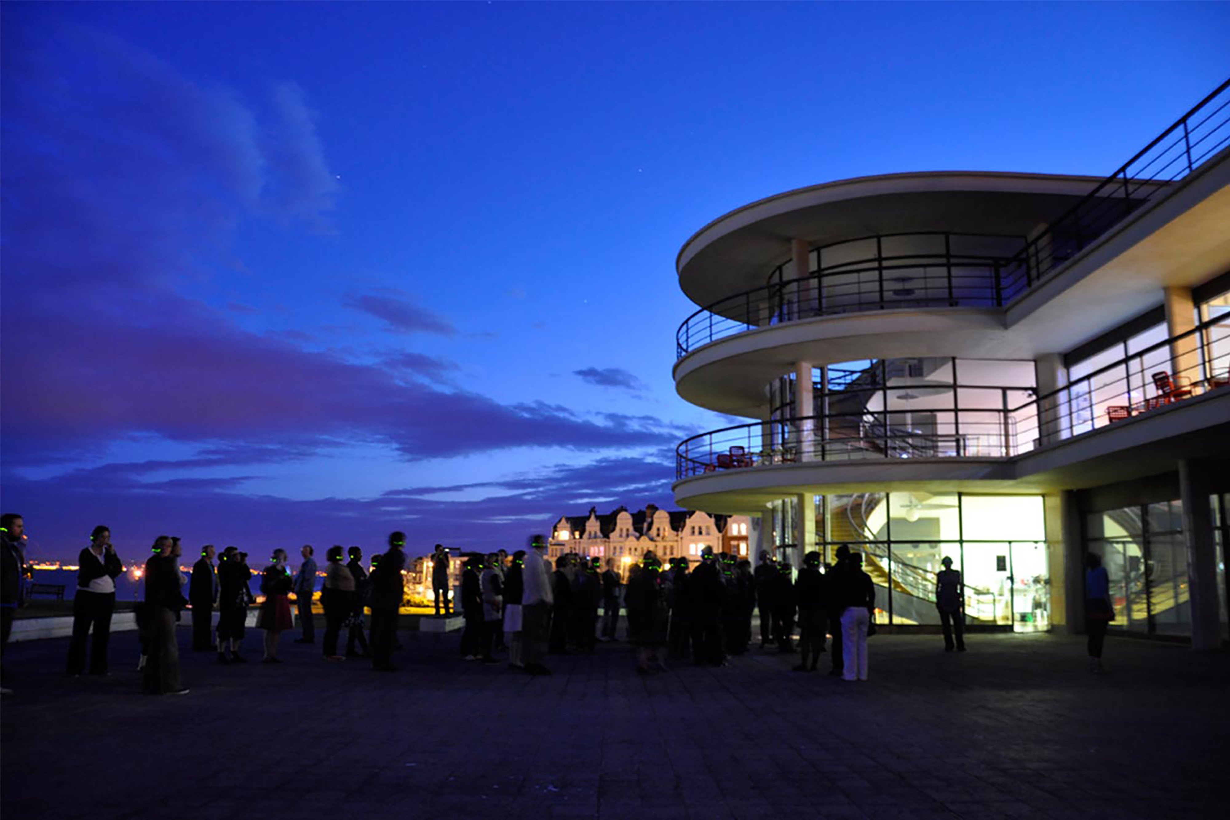 A large white curved building, with light projections around the windows, at night with dark blue sky in the background and an audience watching in rows.