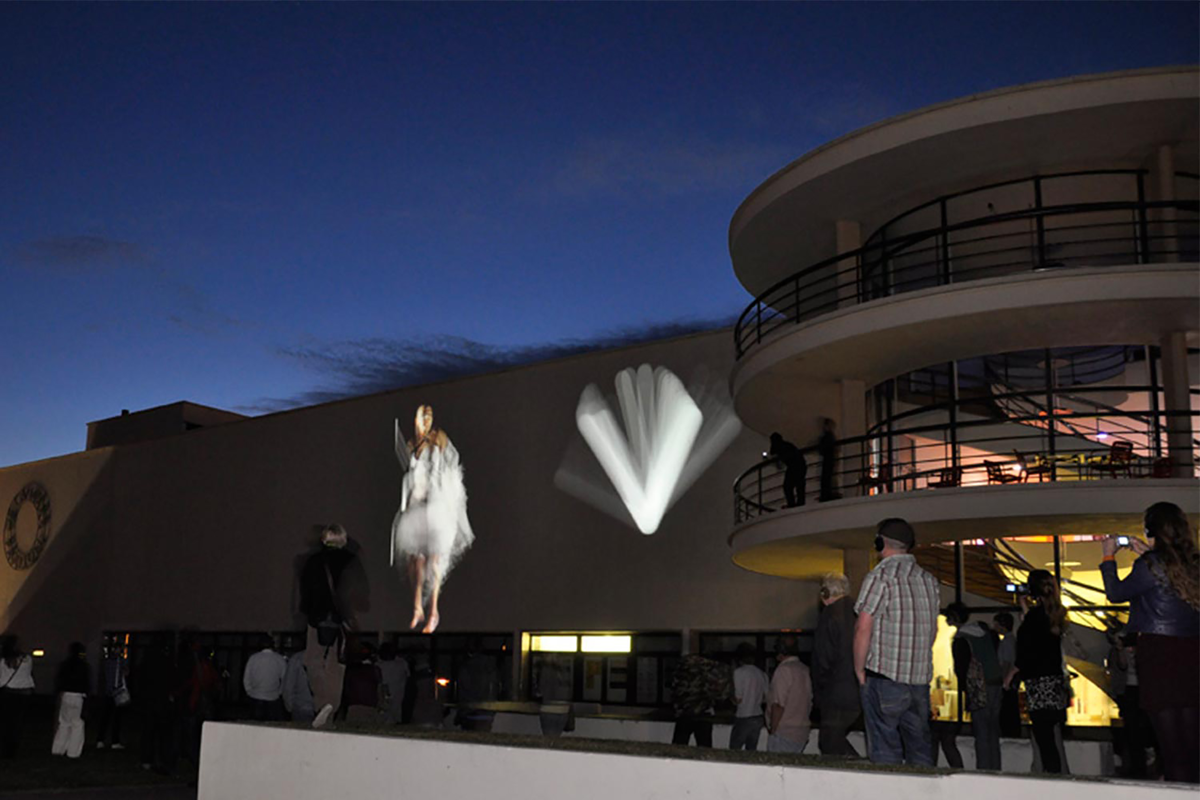 A large white curved building, with light projections around on the walls, at night with dark blue sky in the background and an audience watching .