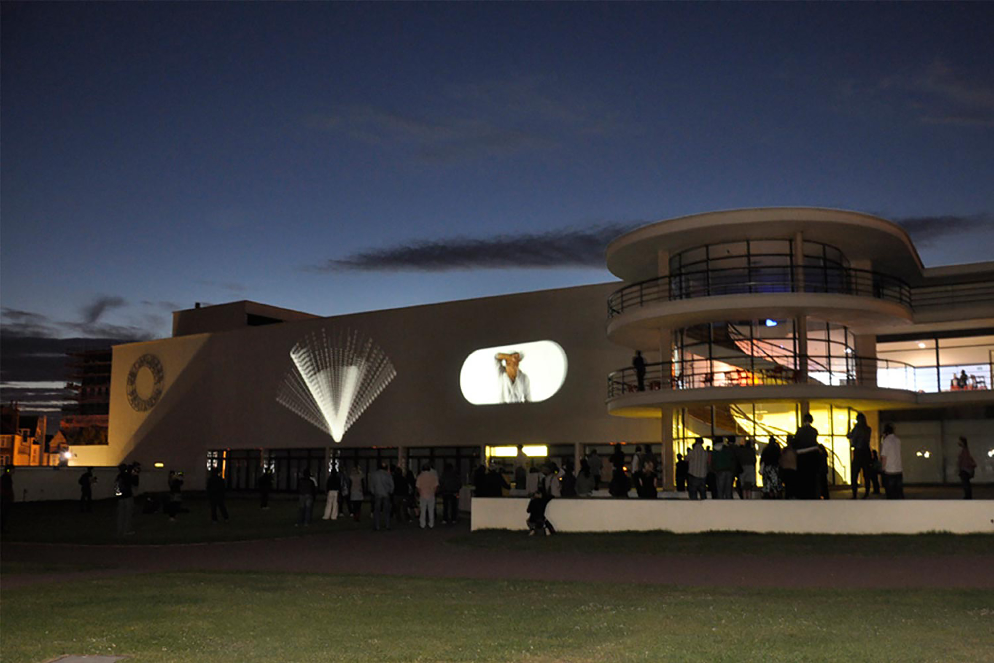 A large white curved building, with light projections around on the walls, at night with dark blue sky in the background and an audience watching .