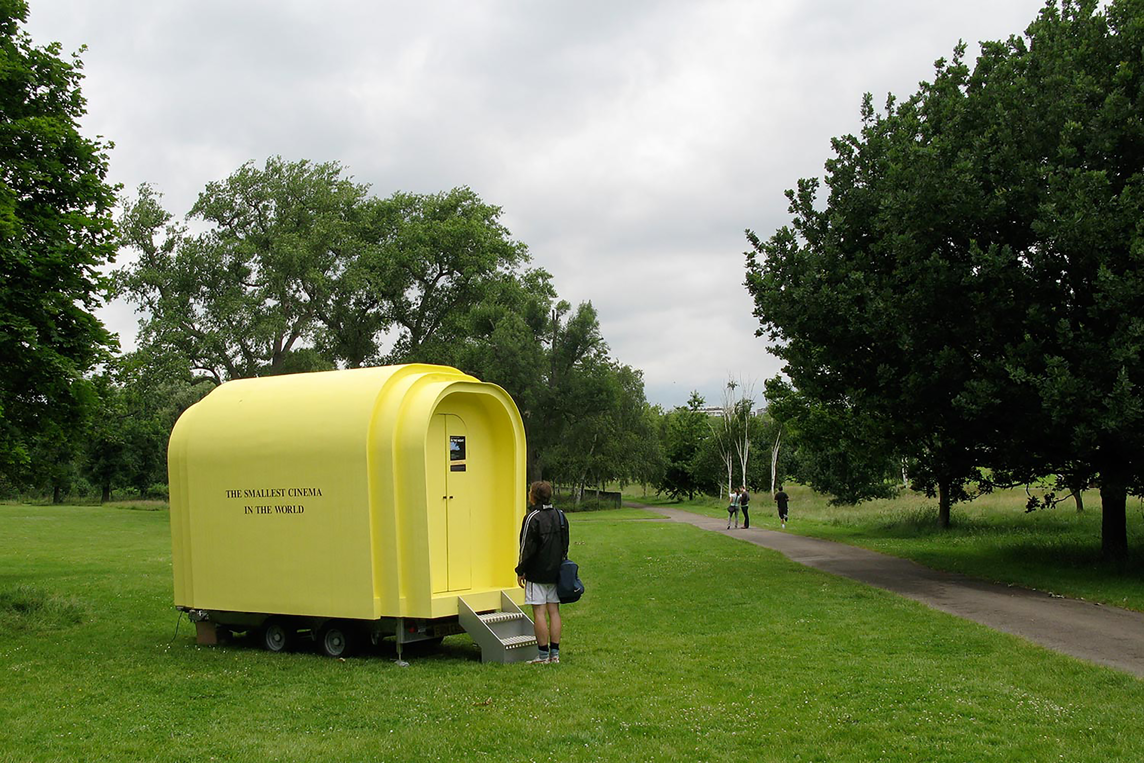 A small yellow outdoor cinema pod with the words "The Smallest Cinema in the world" on the side, with people climbing inside.