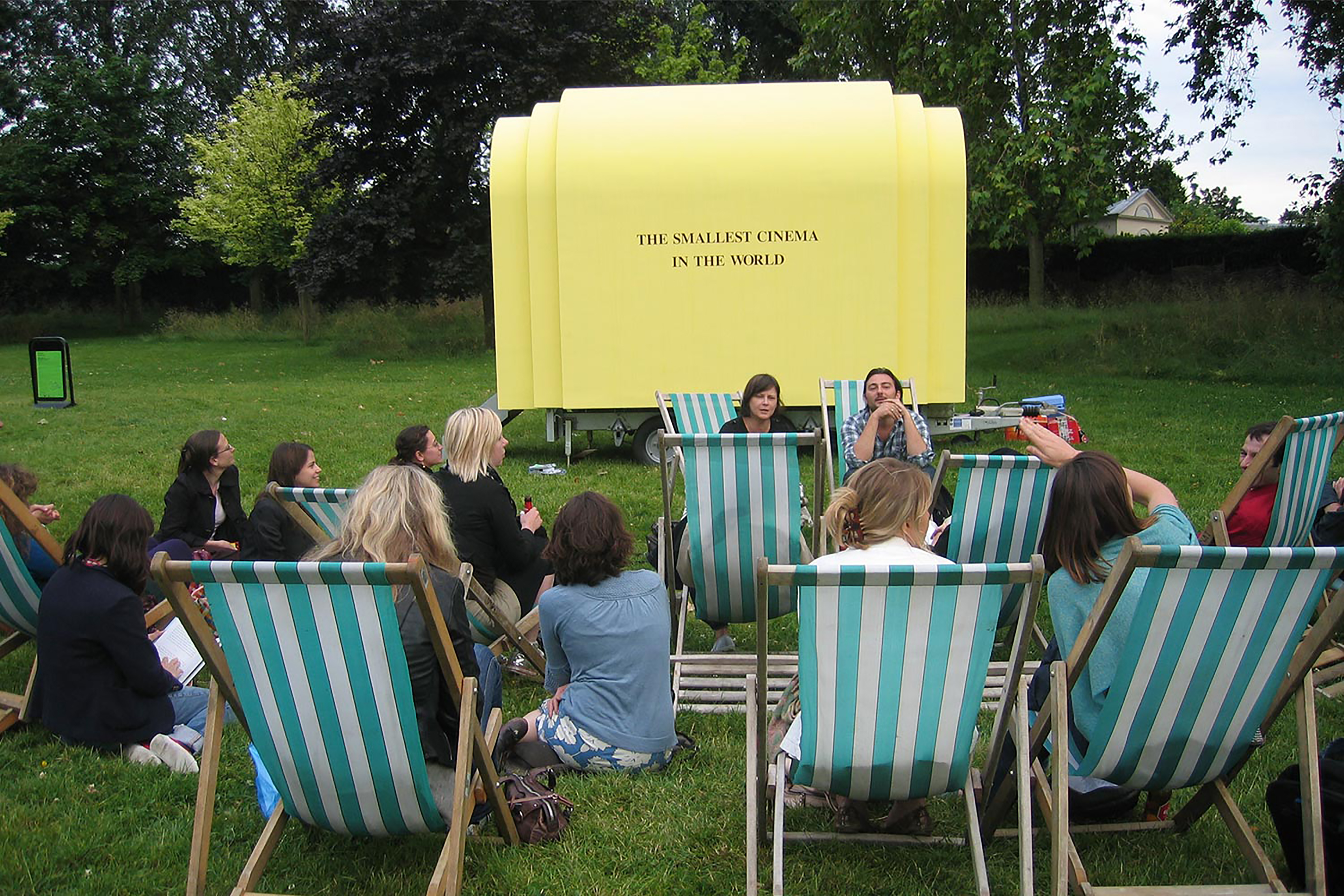 A group of people sitting in green and white striped deck chairs in front of a small yellow outdoor cinema pod with the words "The Smallest Cinema in the world" on the side.