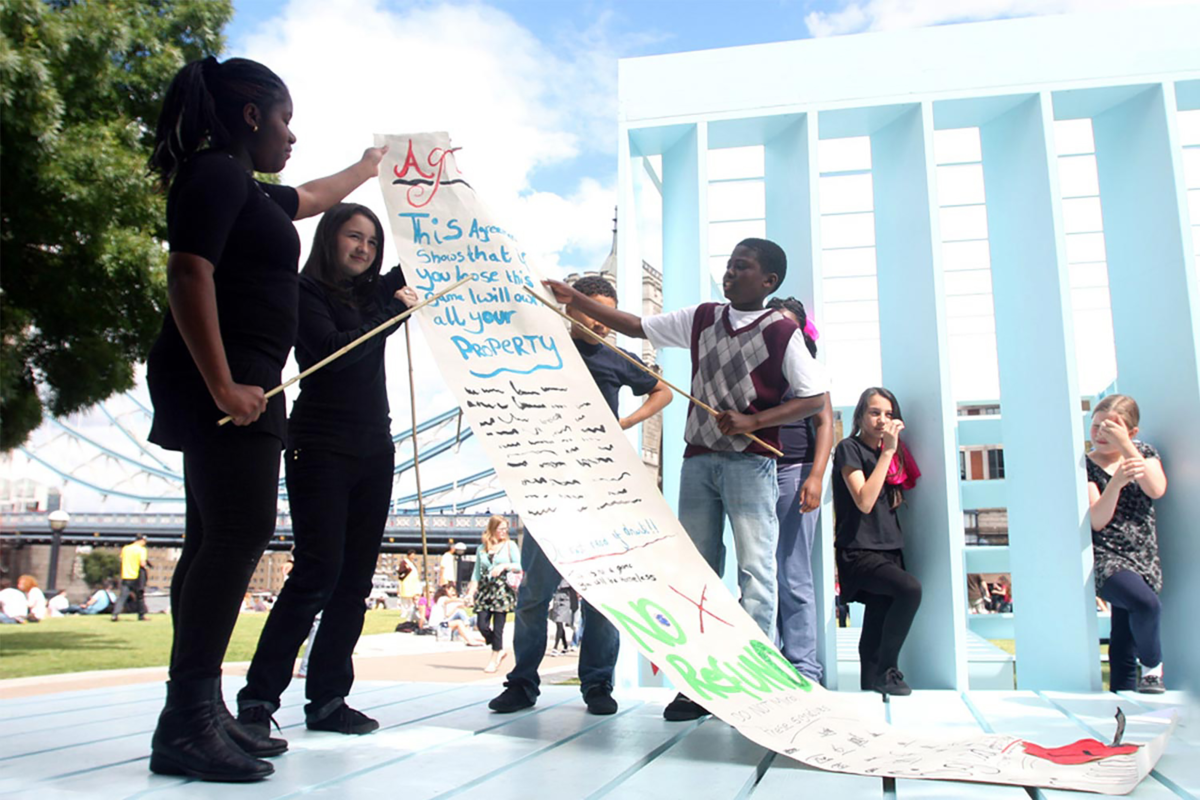 A group of children standing on a powder blue floor next to a wall panel, holding a large roll of paper with handwritten text on it in different colours.