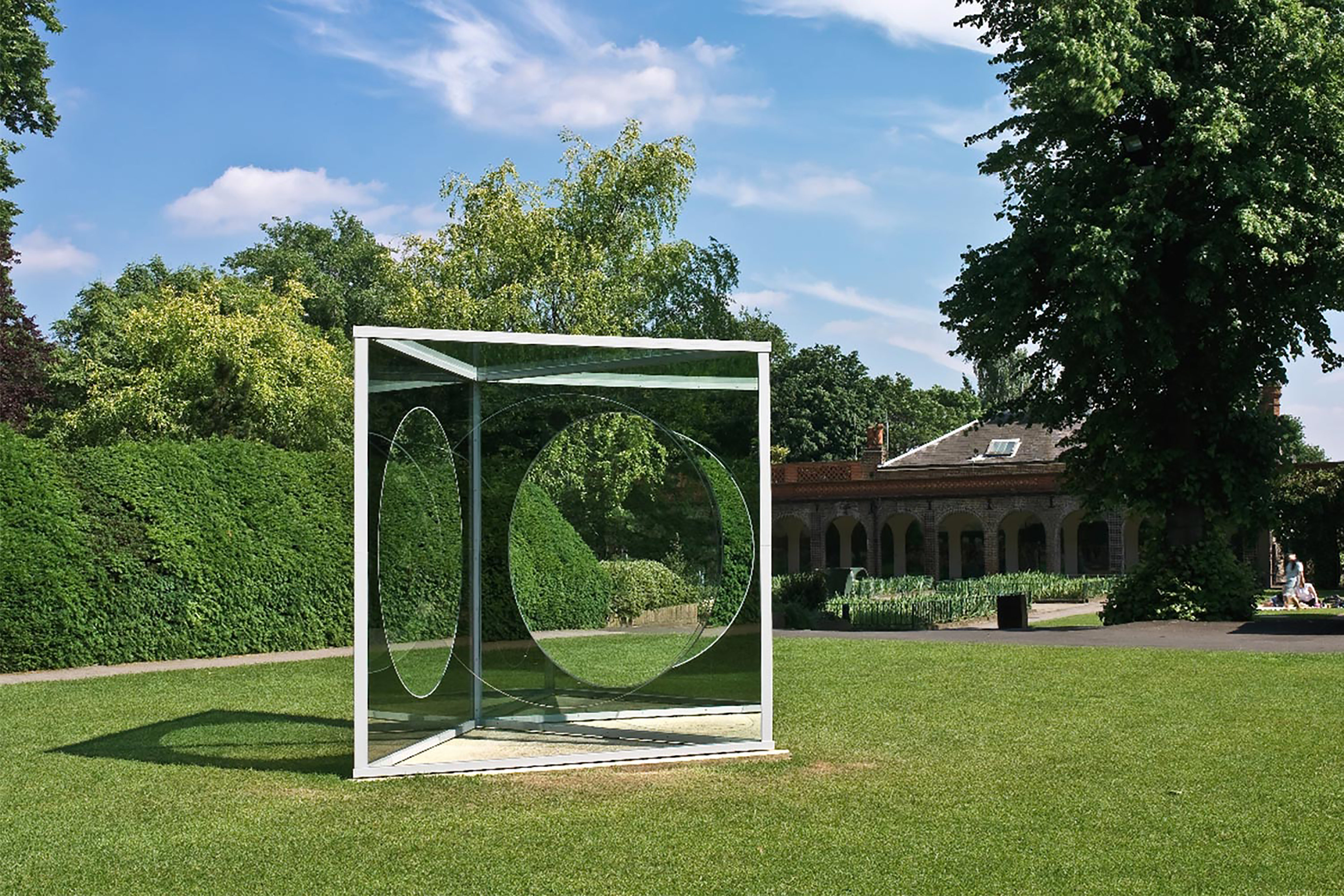 A large triangular sculpture made of reflective material, with large holes cut out, within a green patch of grass.
