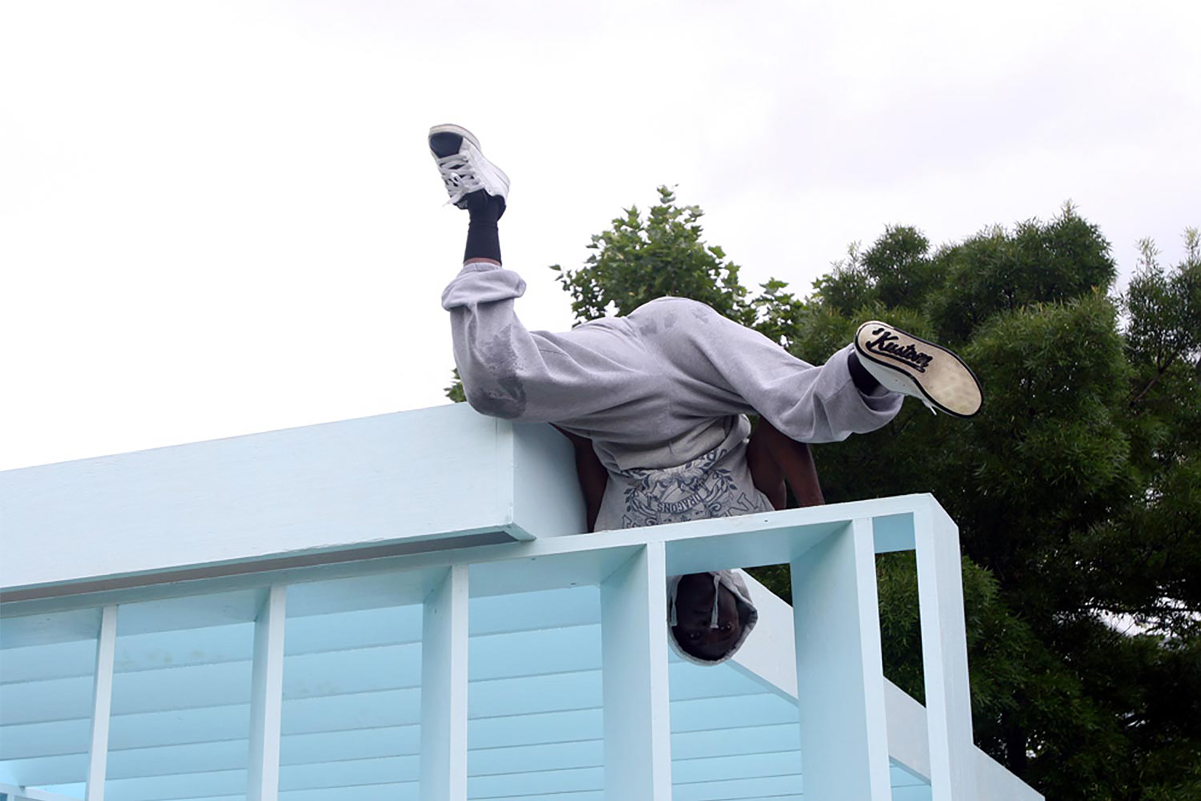 A person climbing on to a powder blue roof over a wall of panels, hanging upside down in a grey tracksuit.