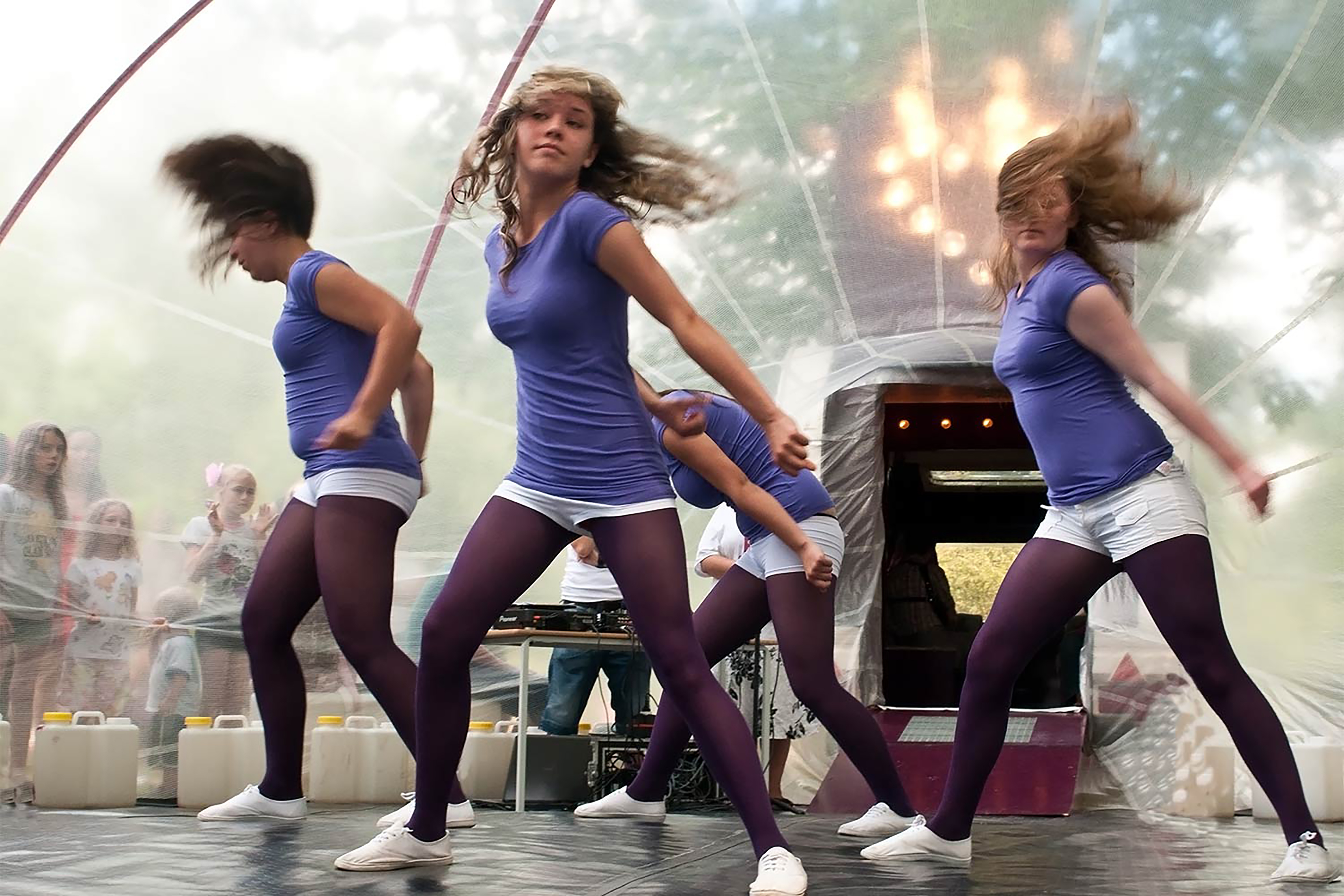 A motion blur photograph of four women dancing inside a transparent tent, all wearing blue tops and white shorts.
