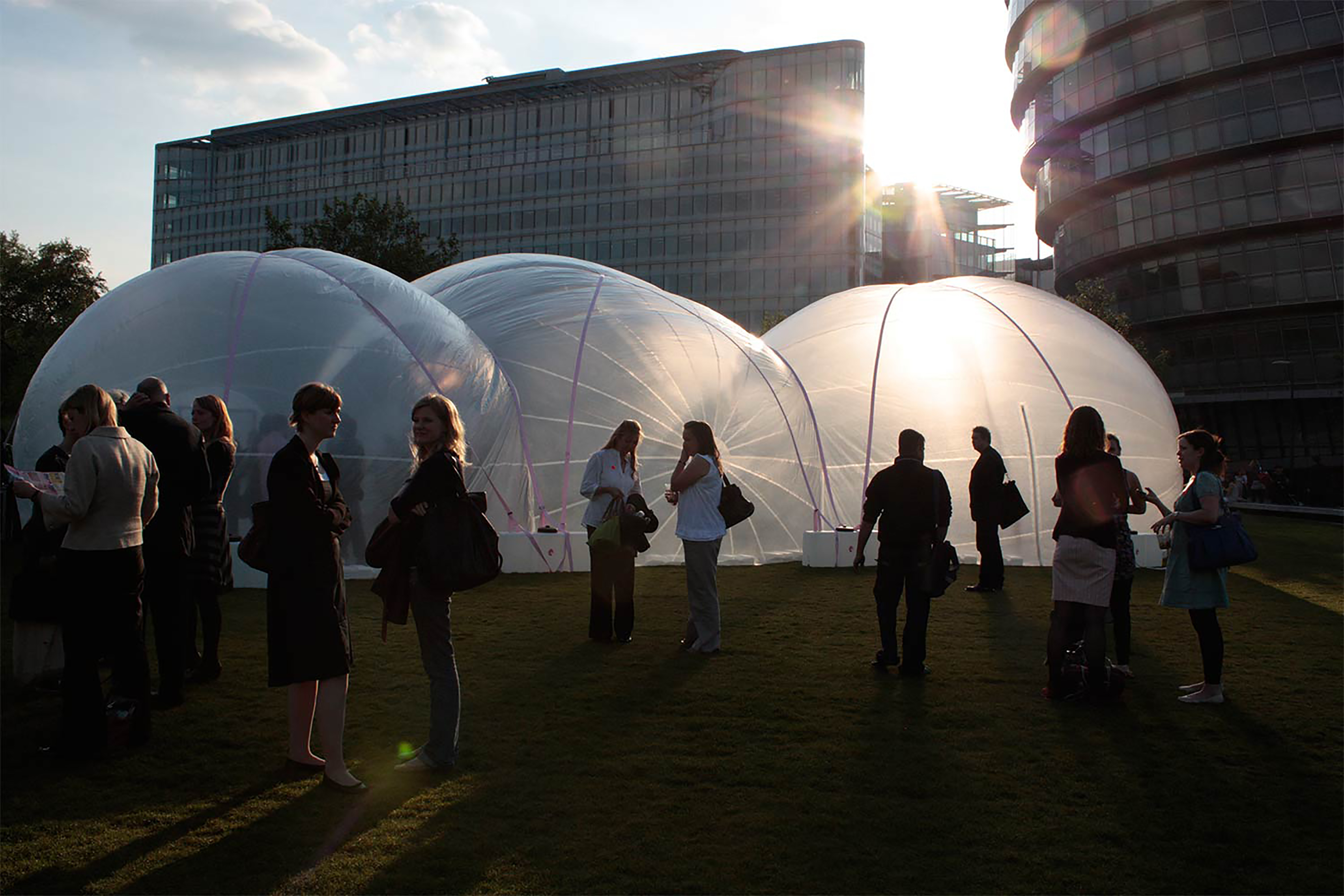 A large see-through plastic inflatable bubble-like structure in a large grass field with a sunset behind a block of flats.