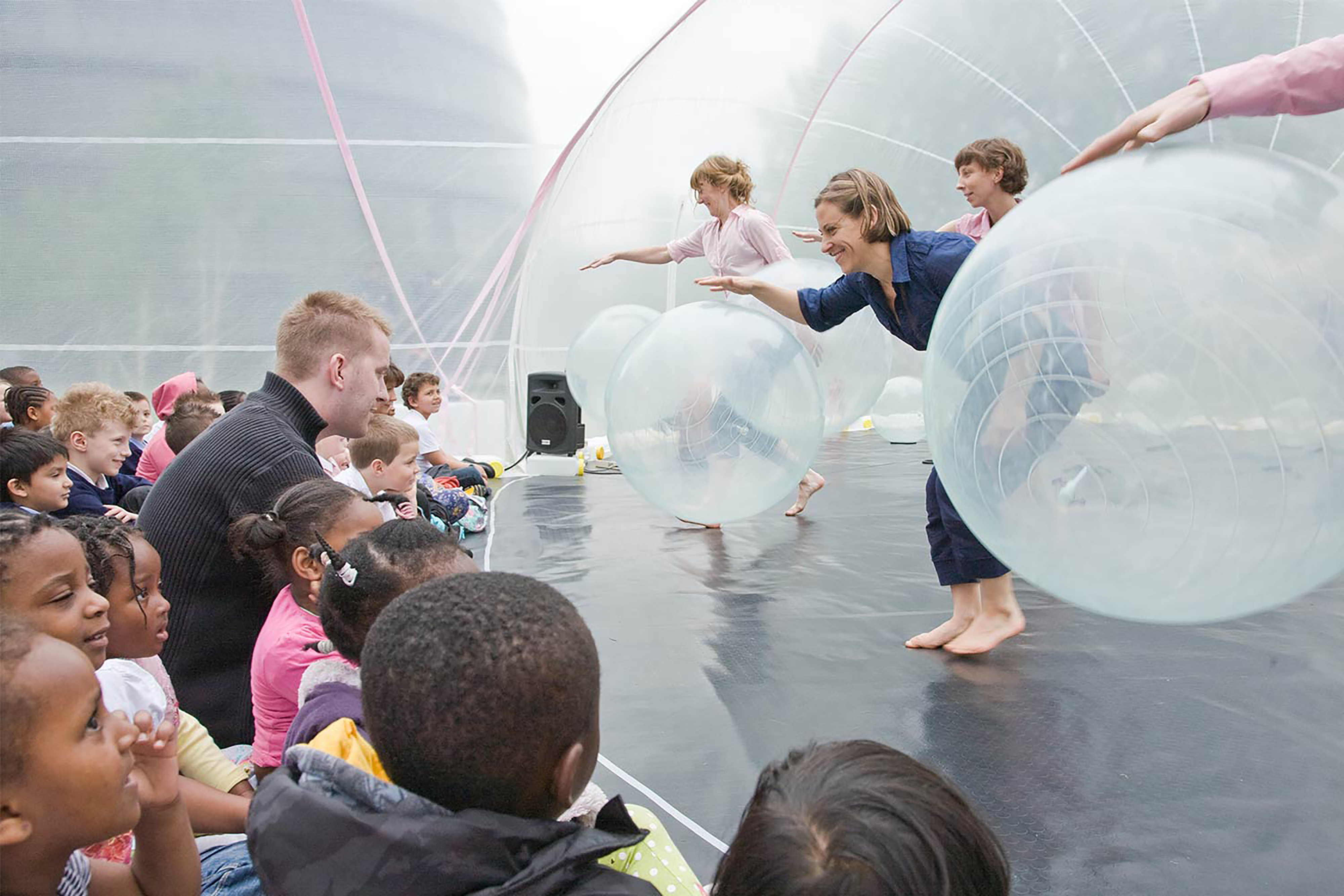 A photograph of three women on stage bouncing transparent gym balls in front of an audience of children in a transparent dome.