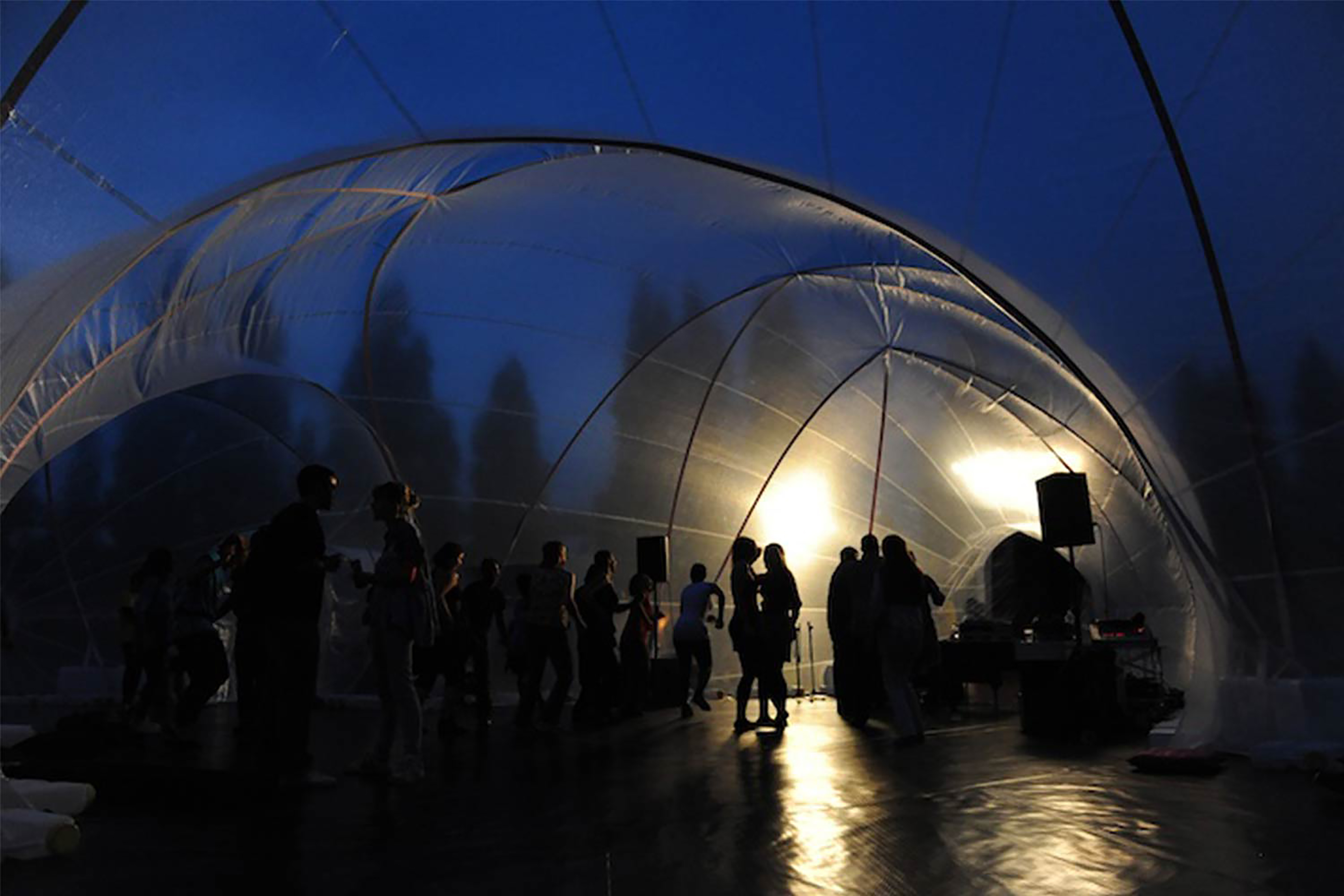 A photograph of a crowd's silhouette against transparent dome tents lit up in the evening time.