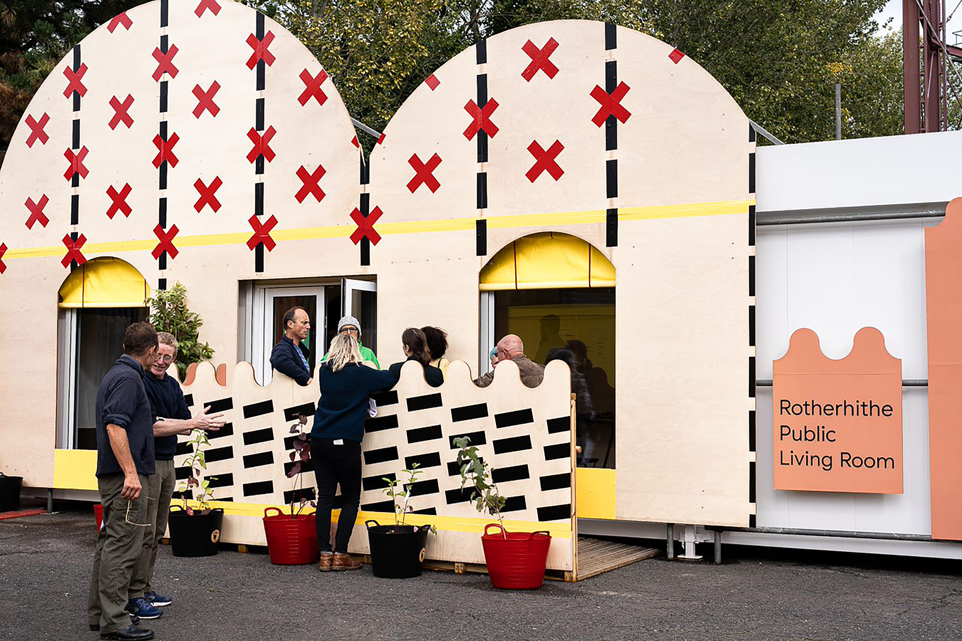 A group of people standing outside a mobile home decorated in red crosses and a pink sign that reads "Rotherhithe Public Living Room".