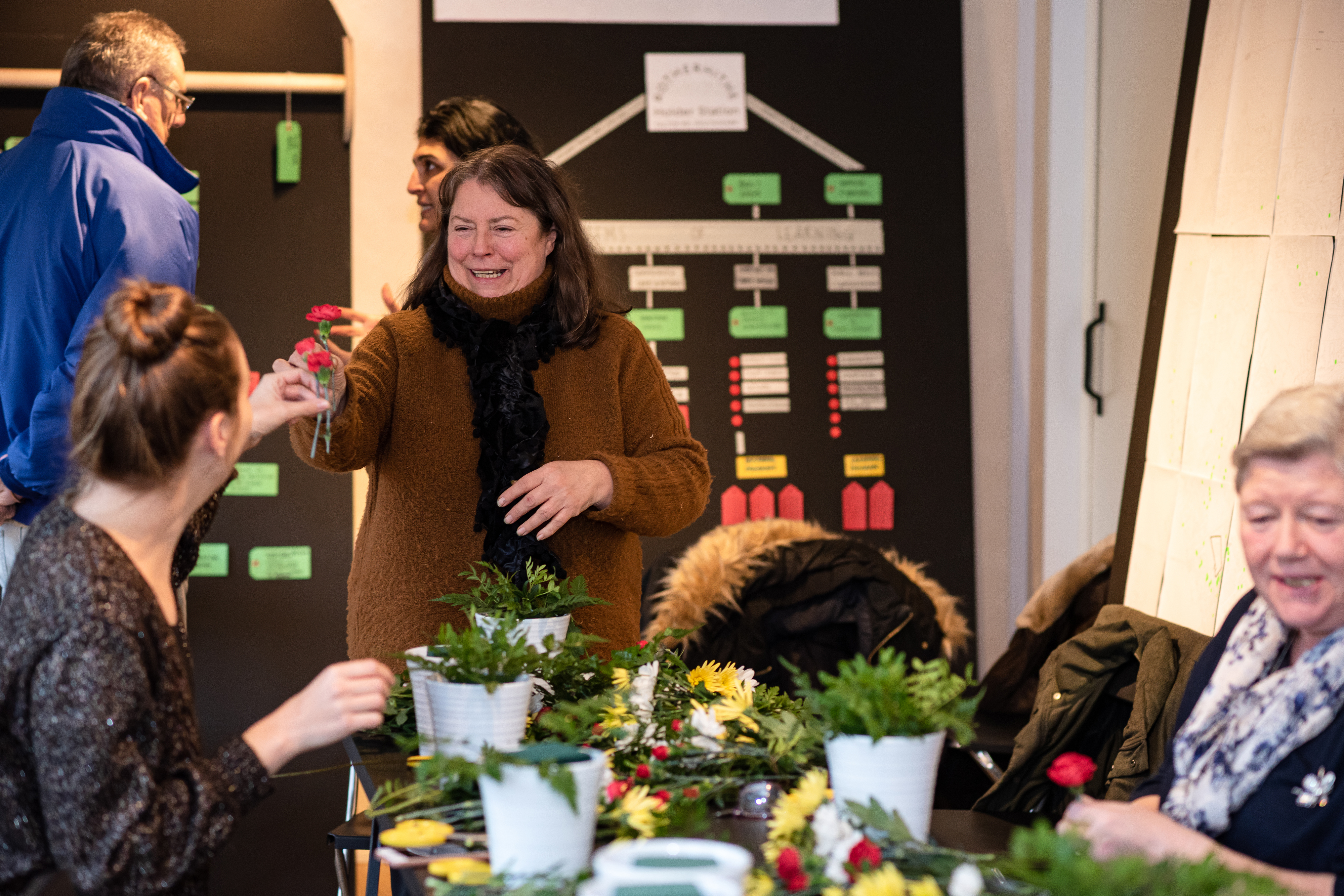 A photograph of a middle-aged woman smiling holding up a flower next to a table covered with flowers and participants inside a mobile home.