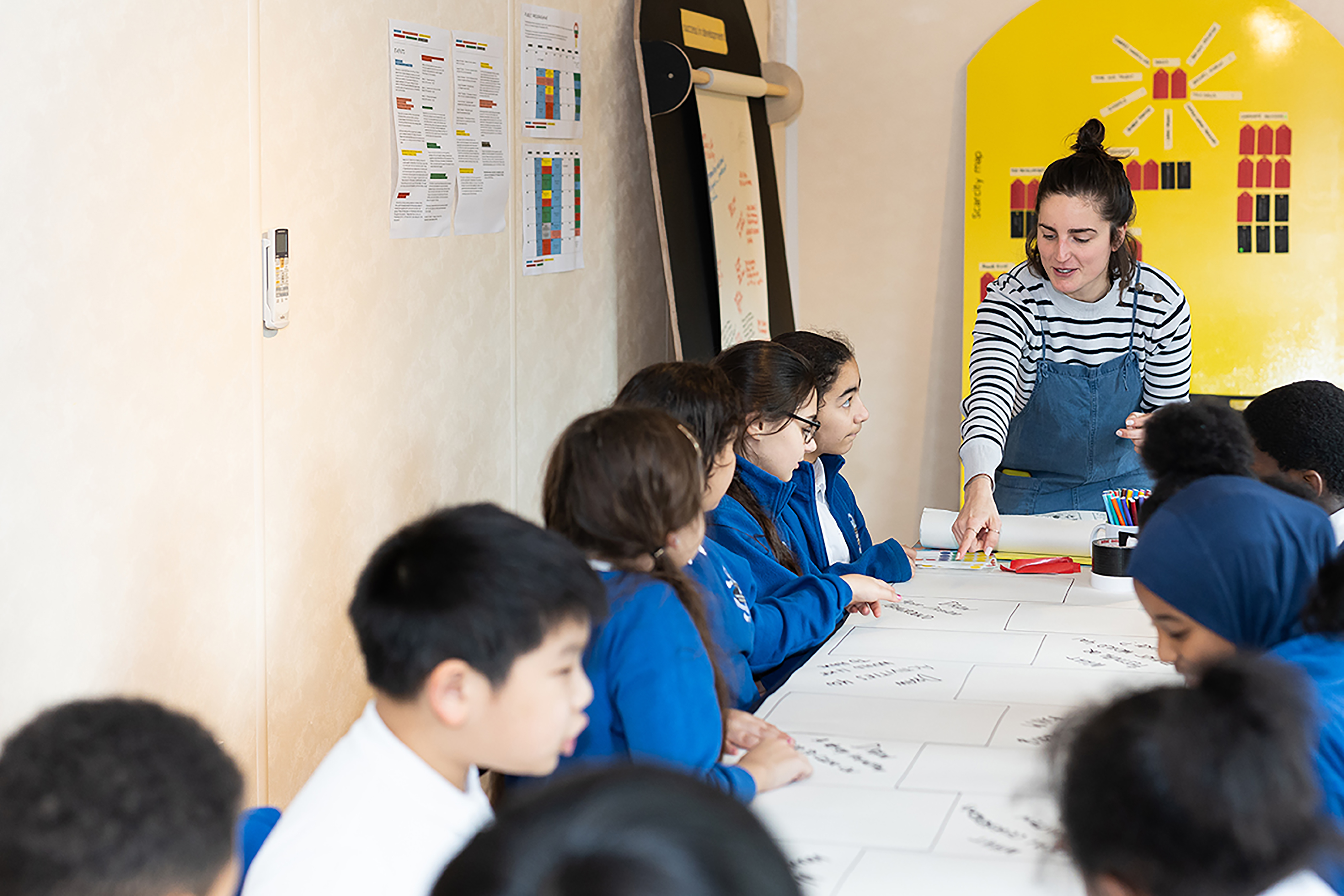 A photograph of a woman wearing blue overalls teaching a class full of children in white and blue tops sat around a table covered with a large roll of paper.