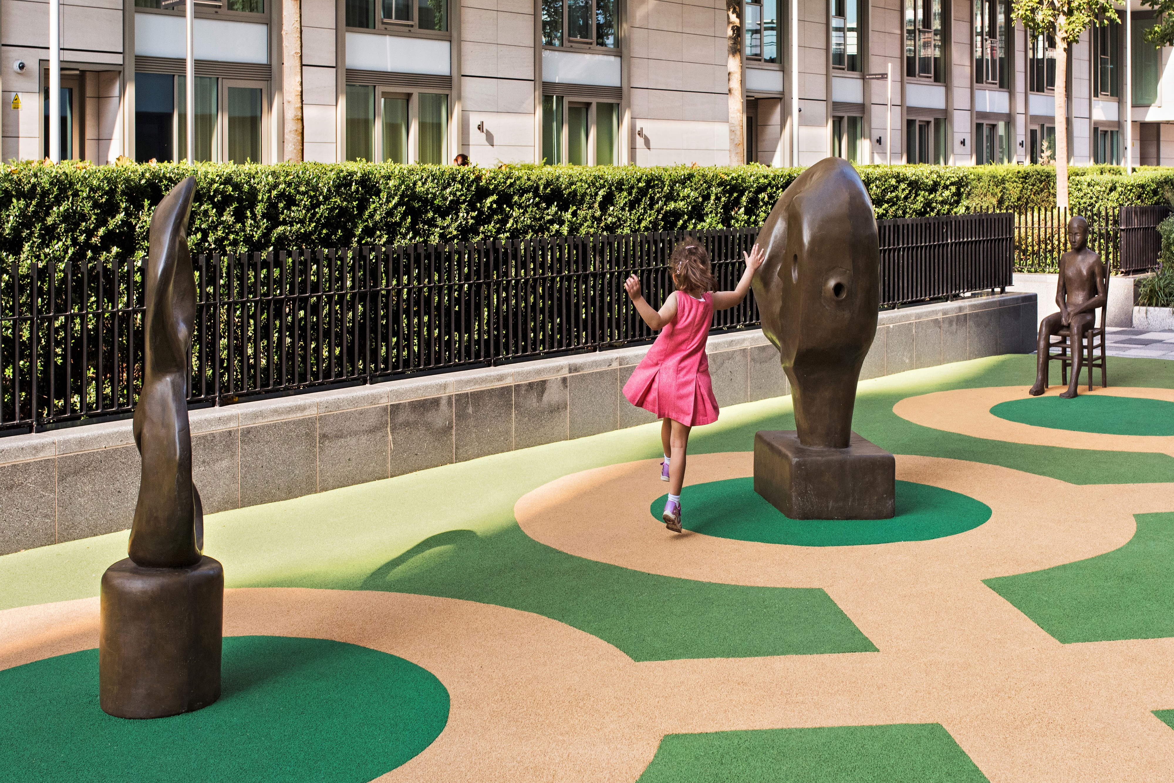 A young girl wearing a pink dress skipping around an abstract bronze sculpture on a green and beige patterned ground inside a public park next to a black metal fence and a large hedge.
