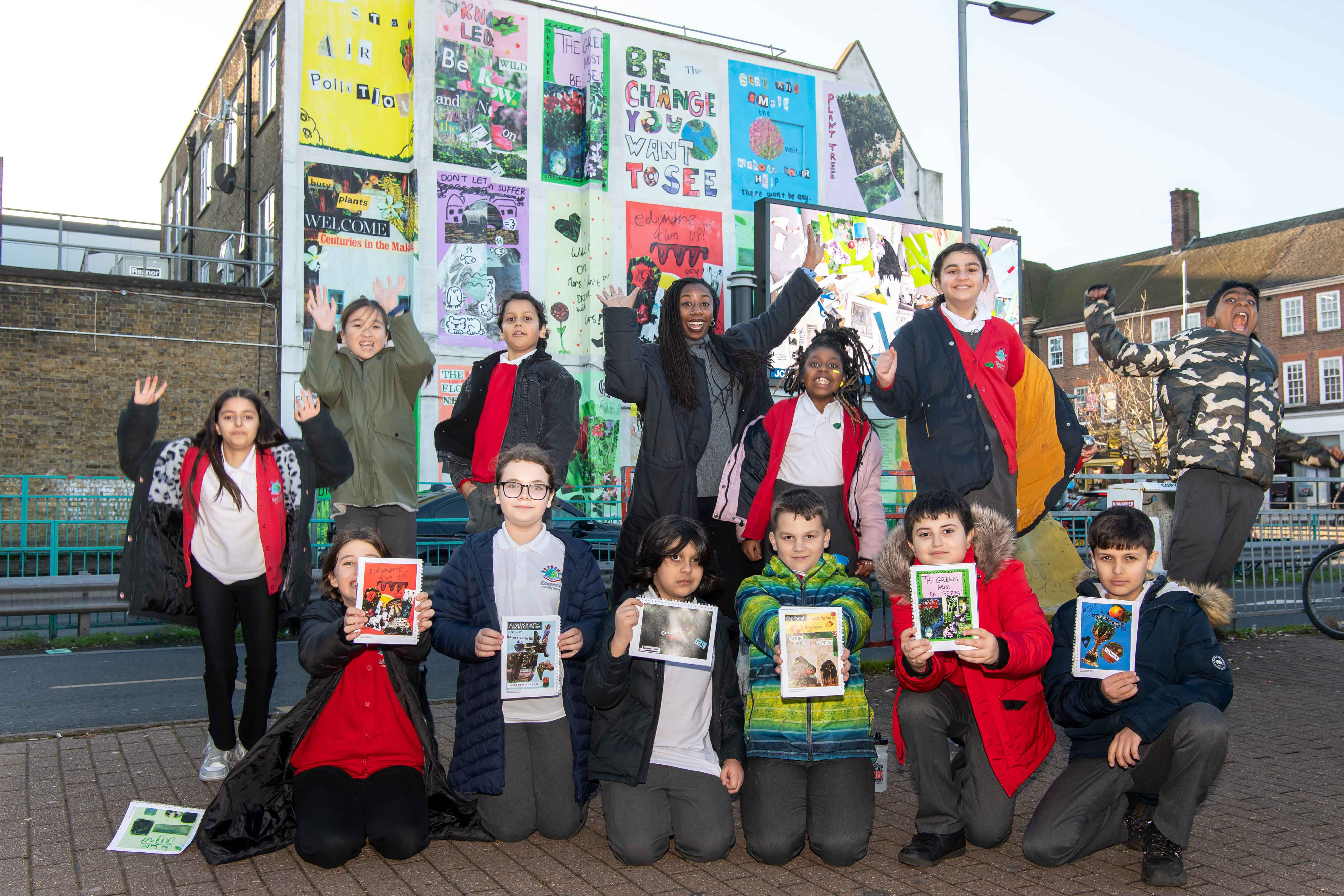 A group of school children and a black woman with long hair. They are all jumping, smiling and holding little booklets with images on. They are standing in front of a large wall covered in big colourful posters.
