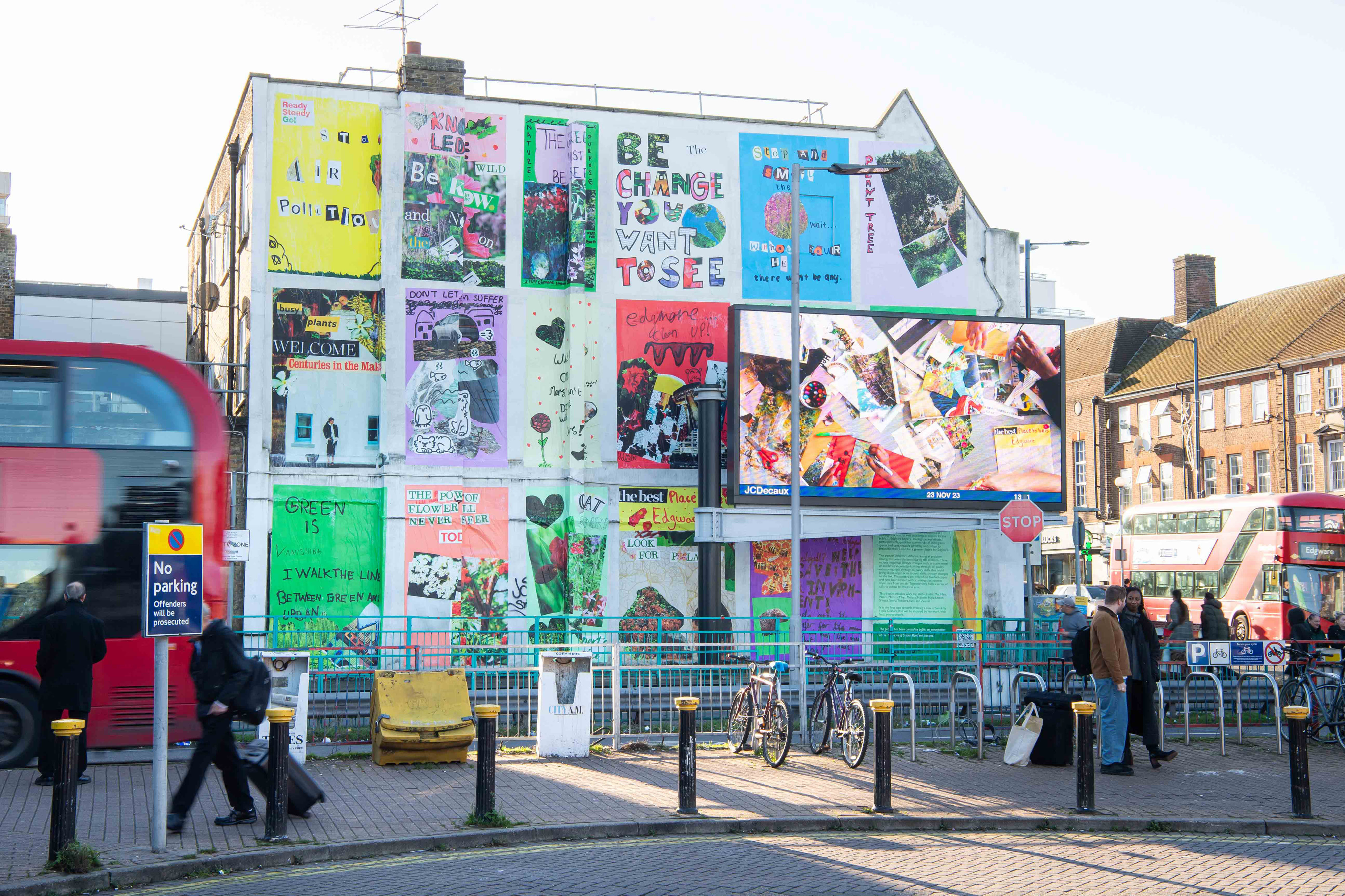 A large wall covered in big colourful posters. In front is a large screen and people walking past.