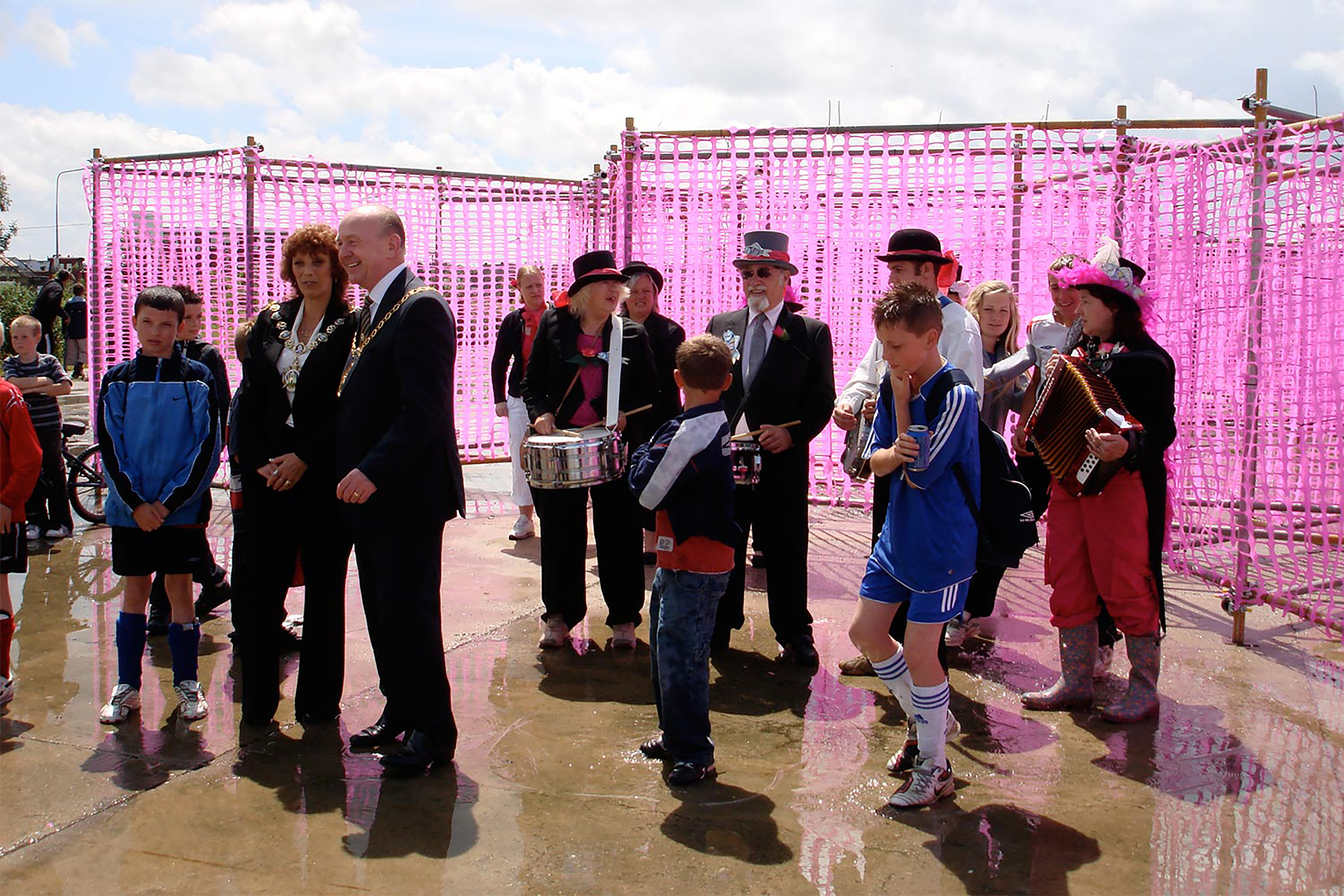 A photograph of a group of adults and children loitering outside a maze constructed of pink tape.