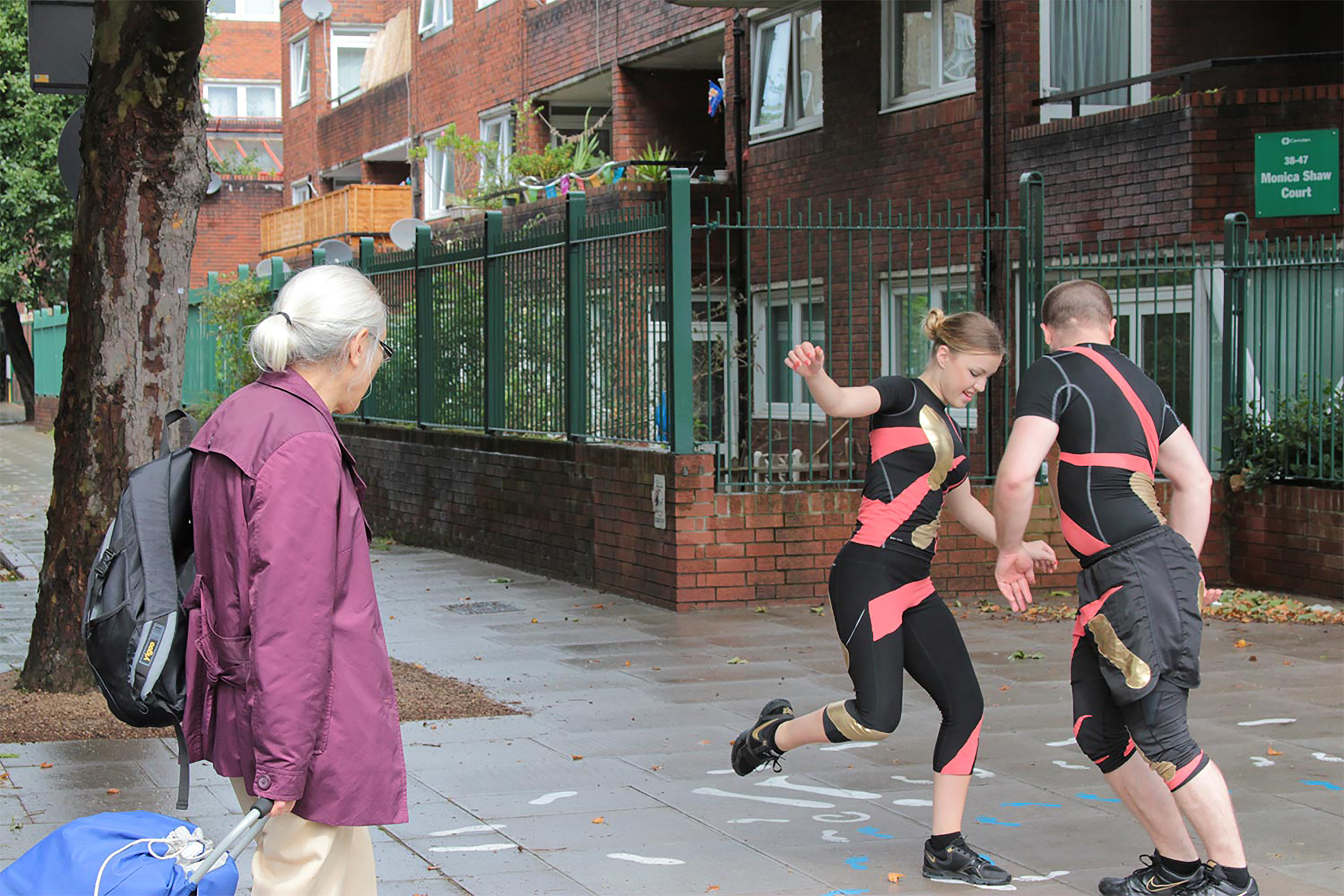A man and a woman both wearing black and pink sporting outfits, dancing outside a red-brick building.