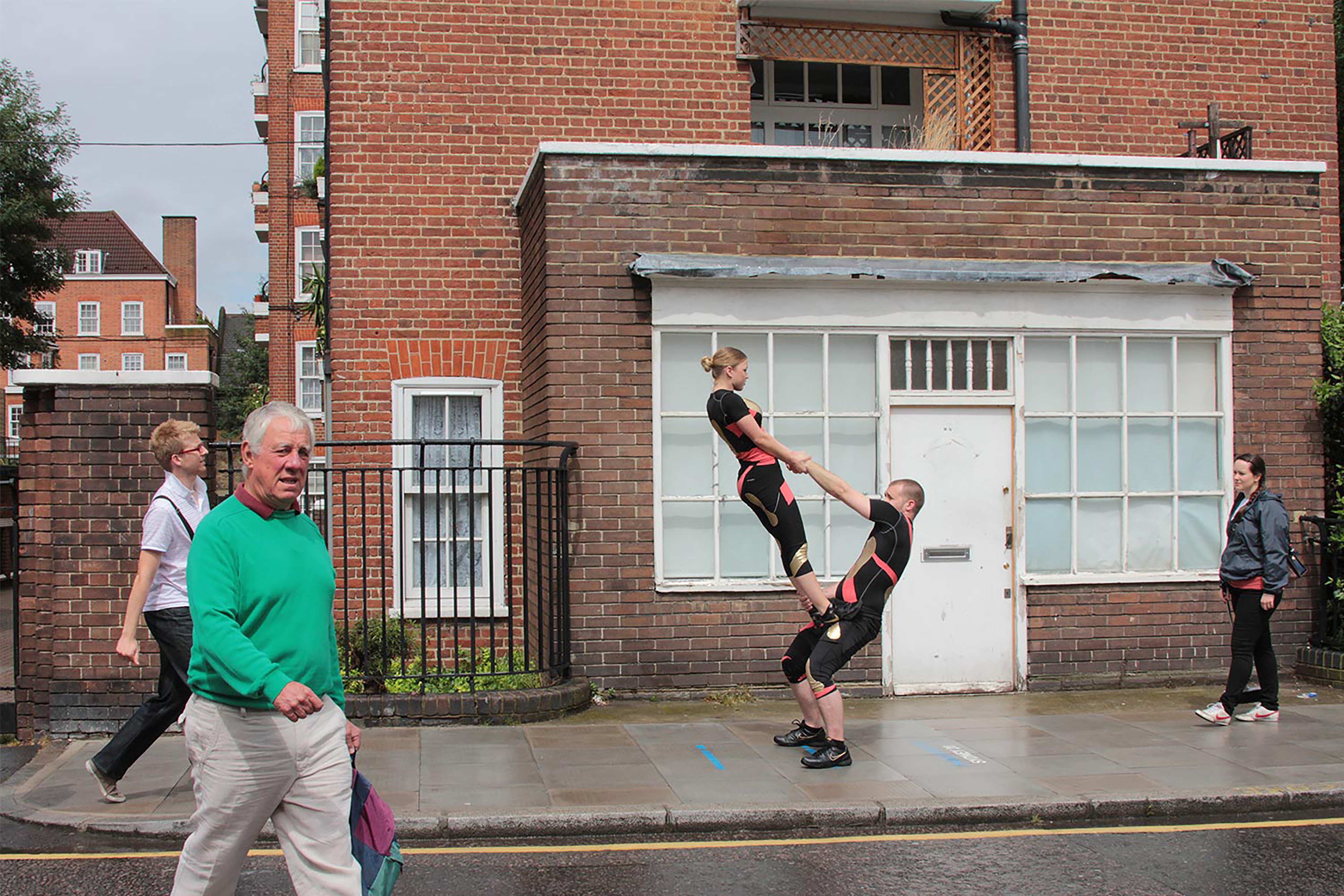 A man and a woman both wearing black and pink sporting outfits, dancing outside a red-brick building with people walking past.