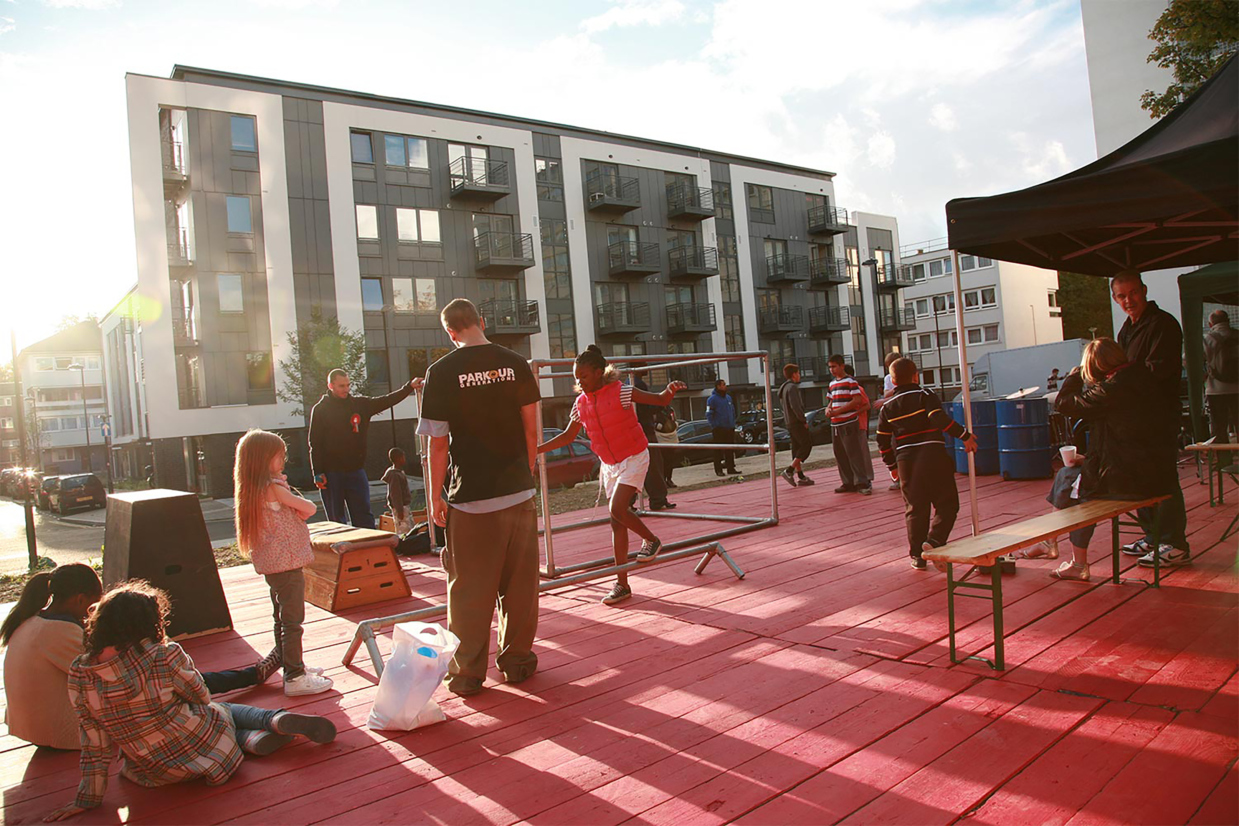 A photograph of children being taught by parkour instructors in an outside training setup deck with red wooden floor.