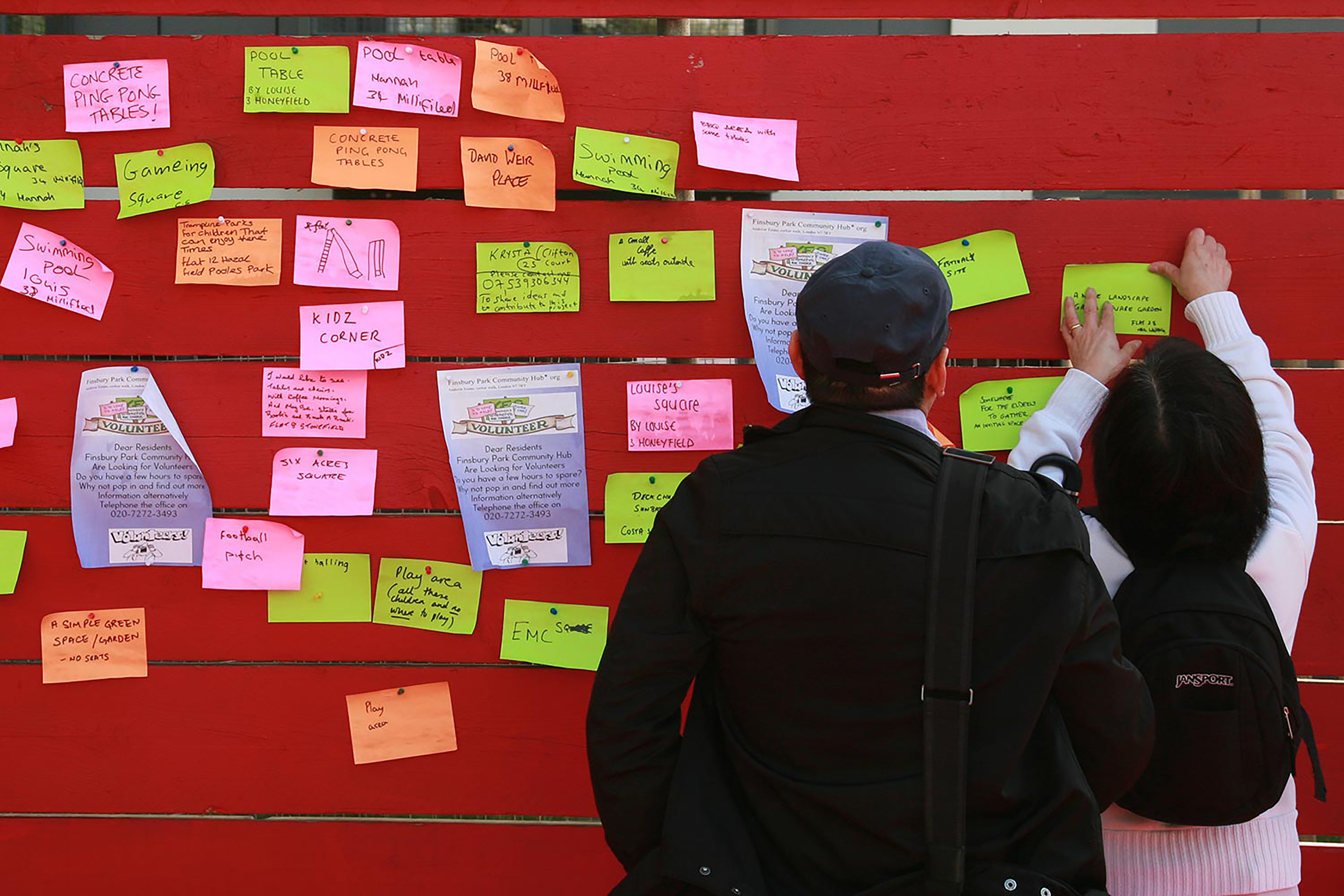 A photograph of a man and a woman putting up a colourful post sticky notes on a red wooden fence covered in post sticks.