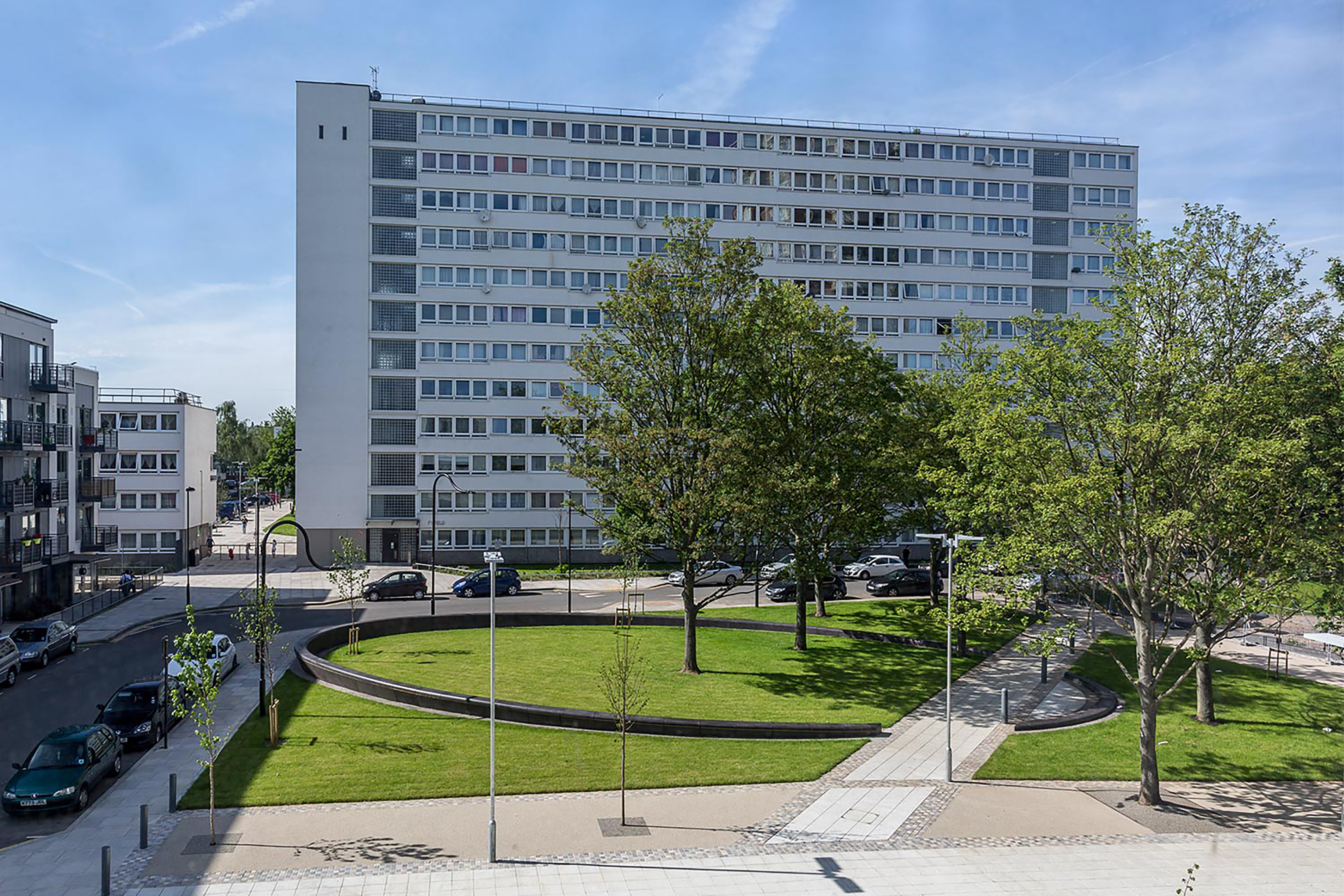 A photograph of a small modern park space surrounded by lampposts and trees in front of an industrial block estate.