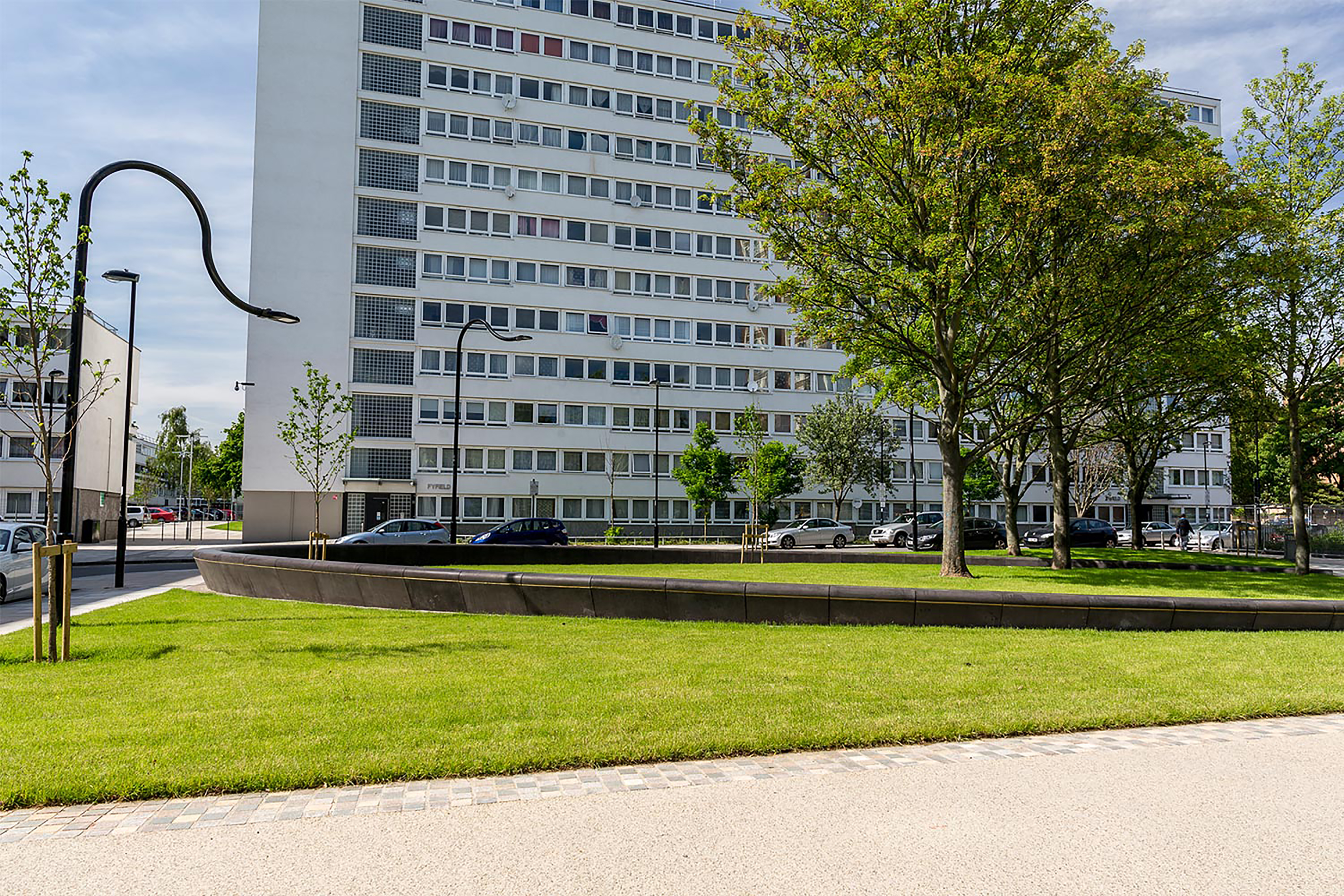 A photograph of a small modern park space surrounded by lampposts and trees in front of an industrial block estate.