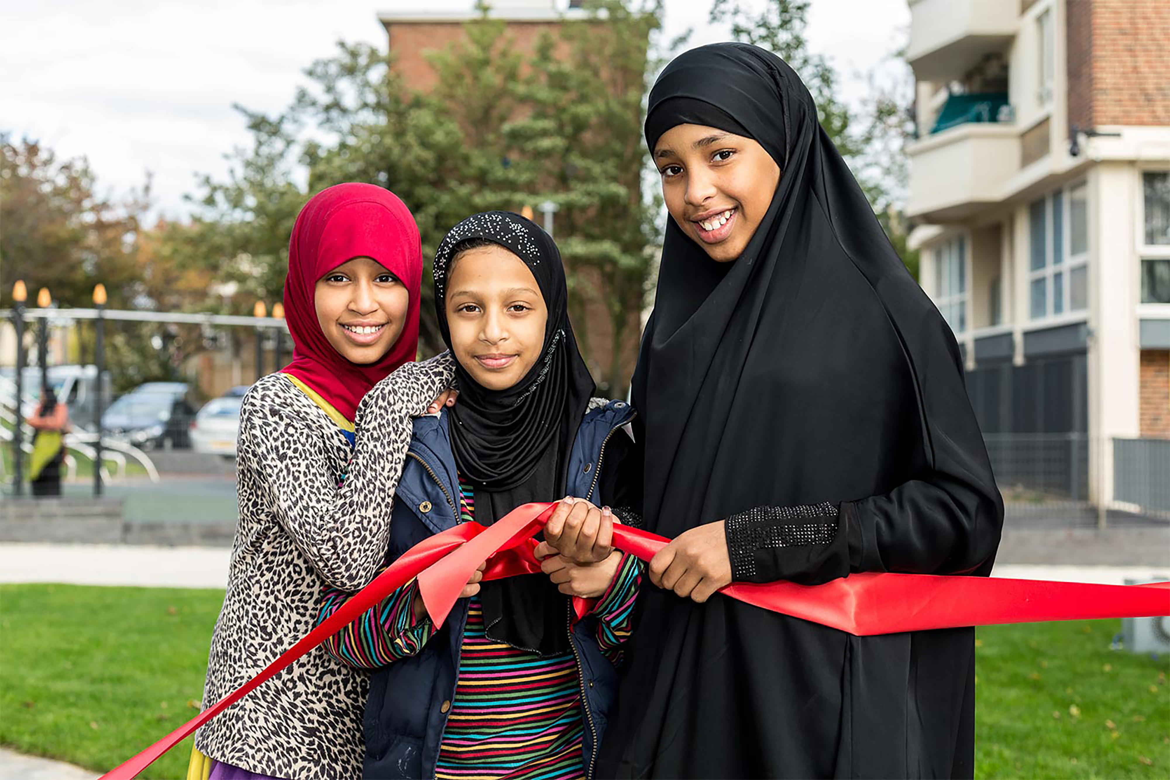 Three young girls wearing hijabs smiling and posing for a photograph while holding a red ribbon in front of an open park space.