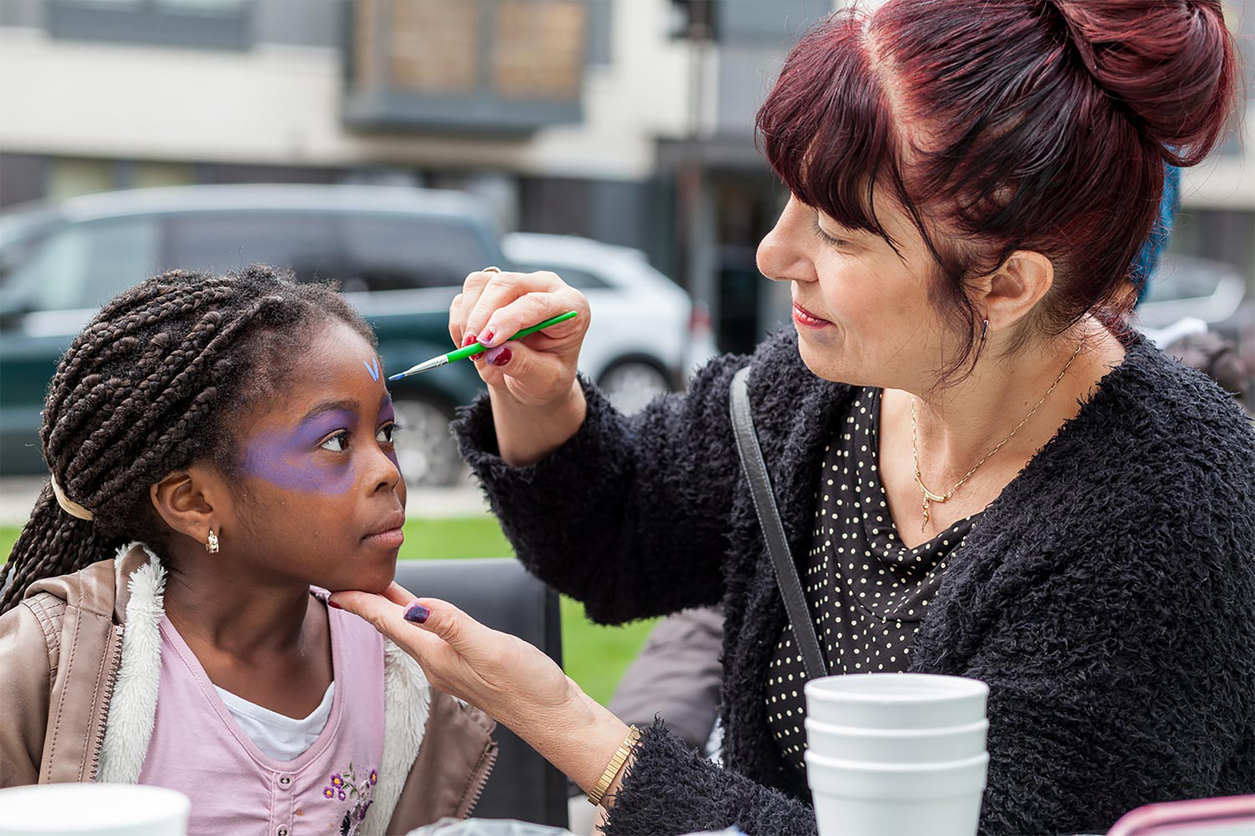 A photograph of a white woman putting face paint on young black girl in front of an open park space.