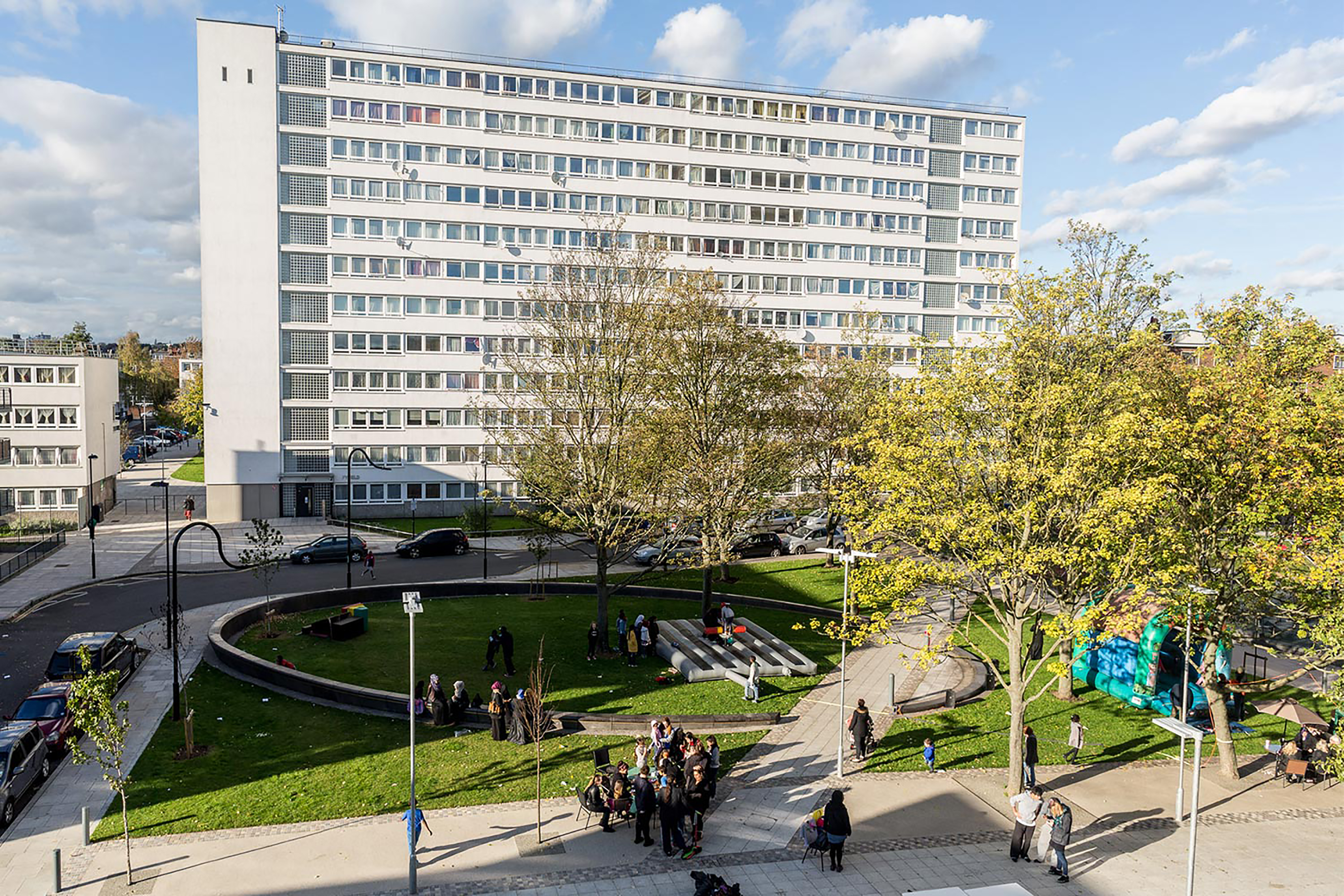 A photograph of a group of school children gathering in a modern park space surrounded by three lampposts in front of a tower block estate.