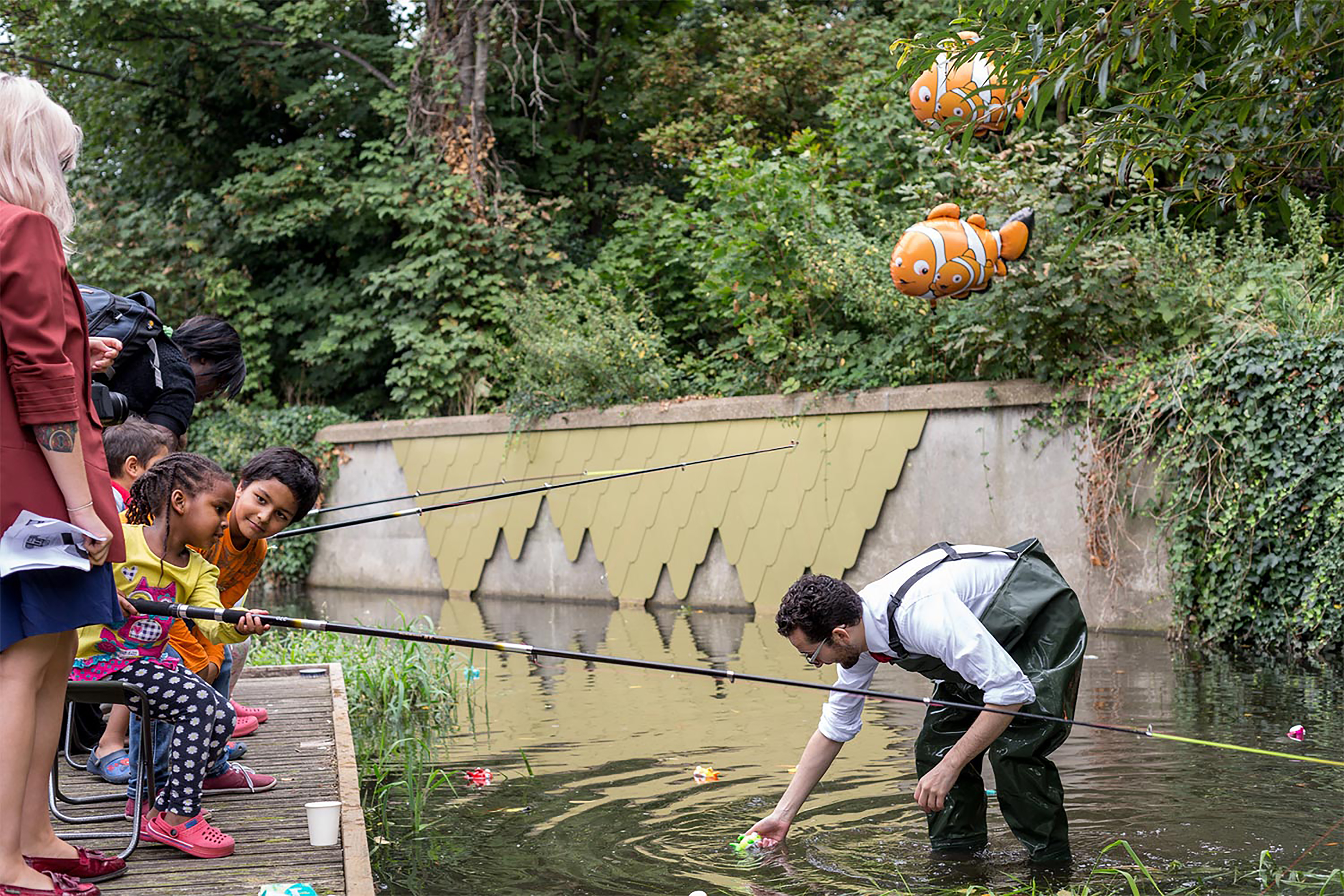 A row of children sitting on the bank of the river, all with fishing rods with a man with fishing waders looks for fish in front of floating clown fish helium balloon.