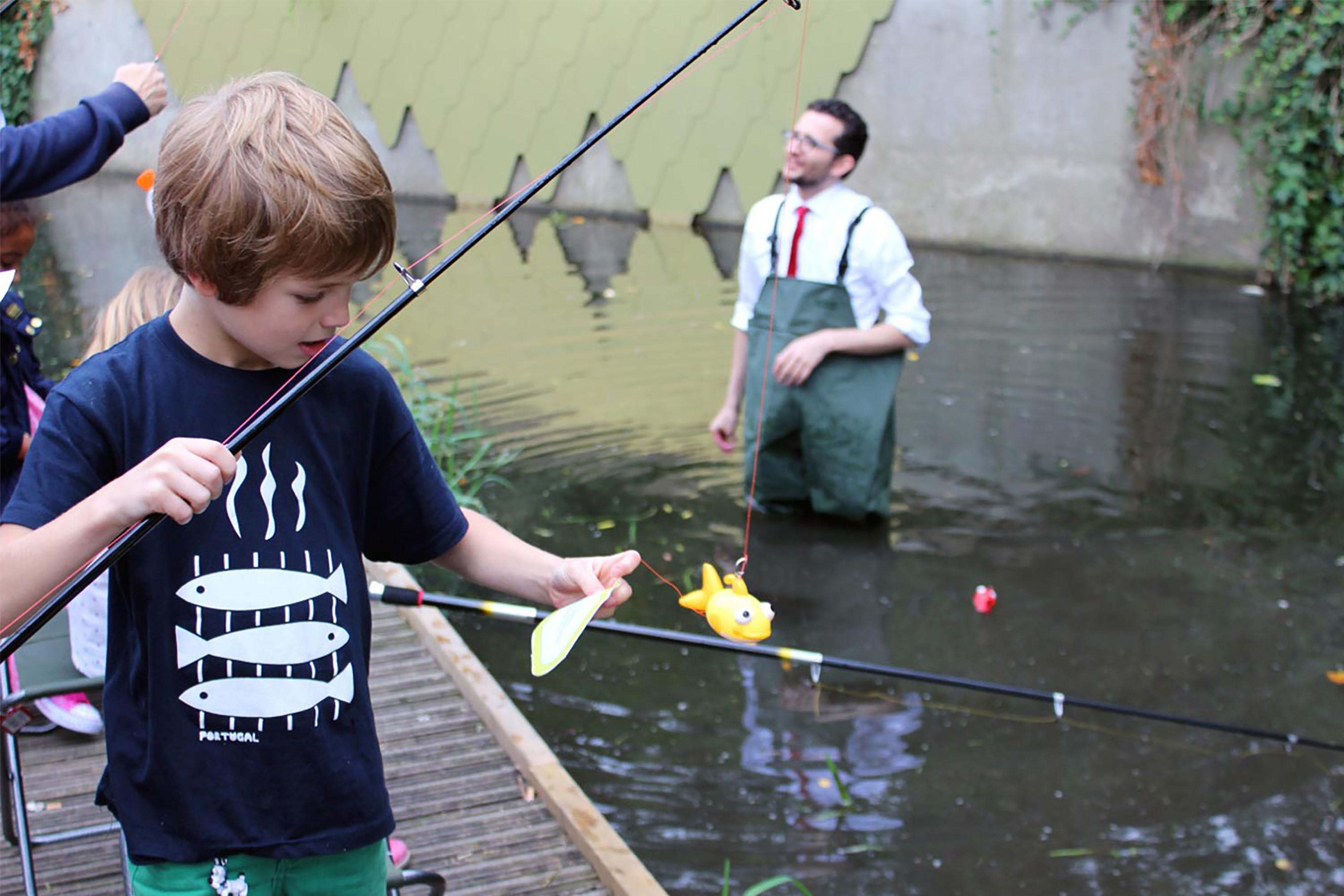 A young boy holding a fishing rod with a plastic fish attached to the end in front of a shallow river.