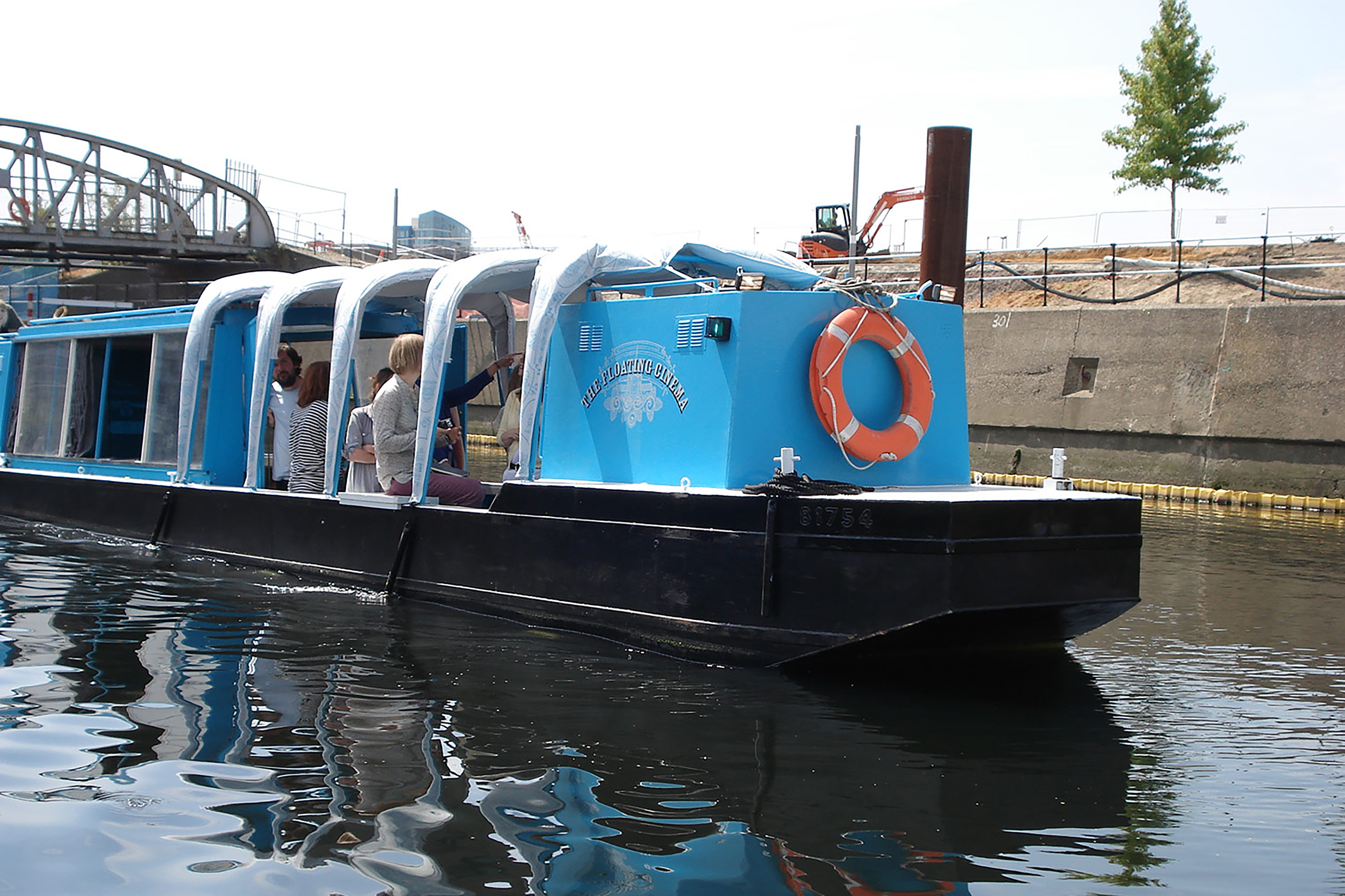 A large blue barge canal boat going down a river with an orange float on the side, carrying a group of passengers, on a bank next to a grey-brick wall.