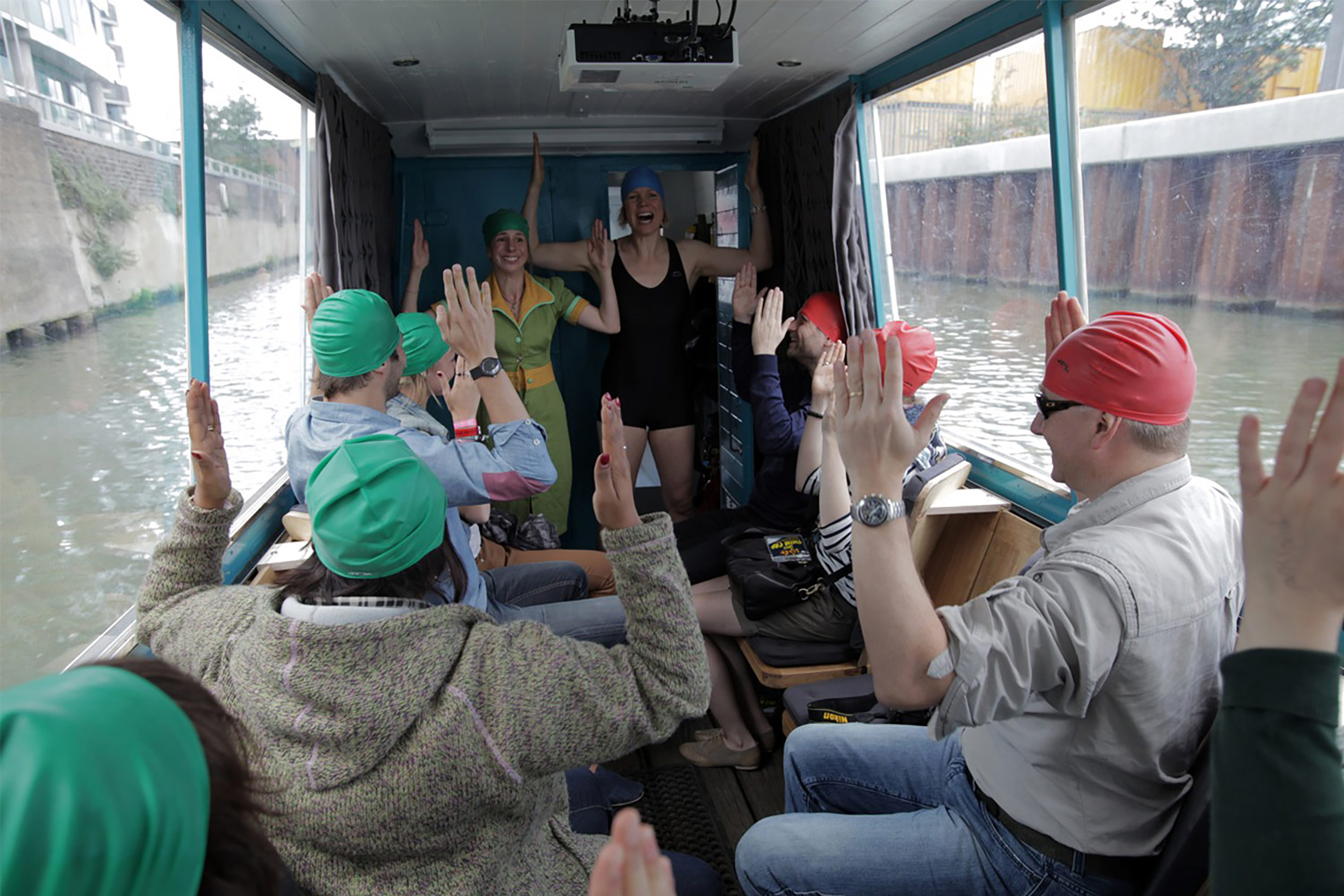 The inside of a barge canal boat carrying a group of passengers, all wearing green and orange swim caps and all holding their arms up smiling.