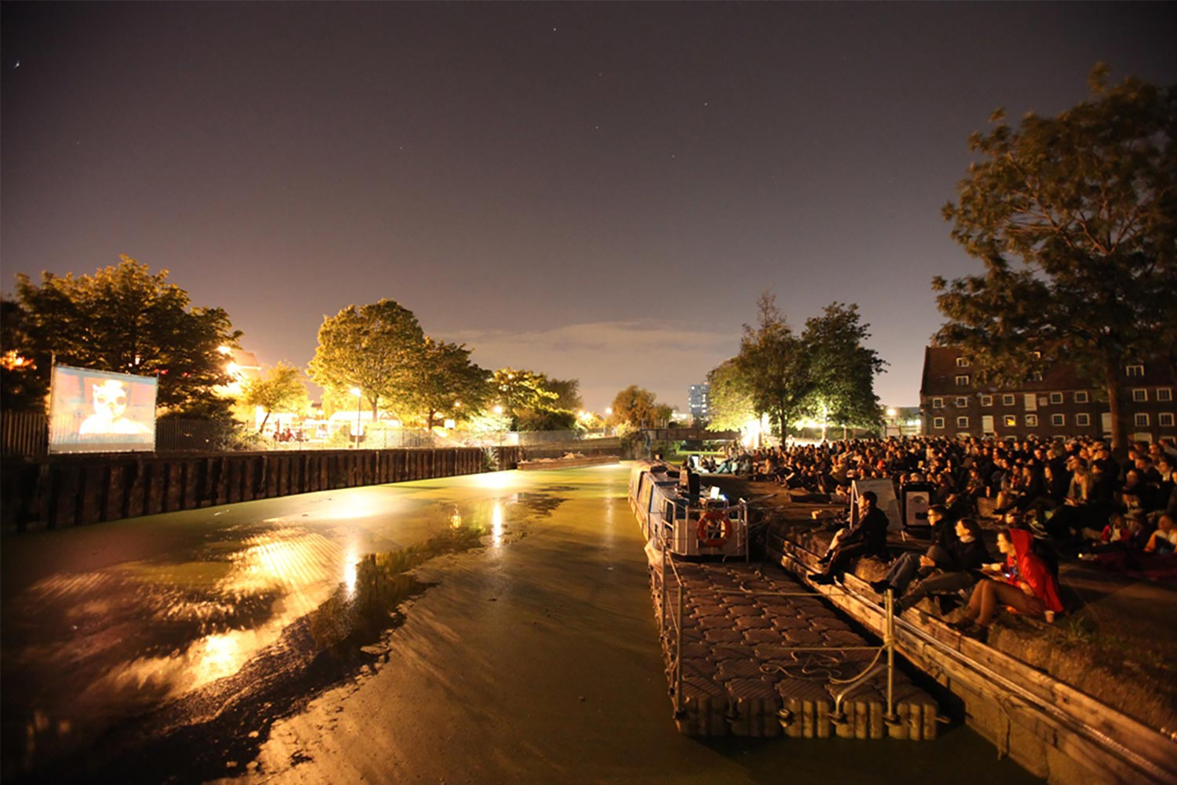 A large blue barge canal on a bank next to a large group of people at night with hanging lights all surrounding the river.