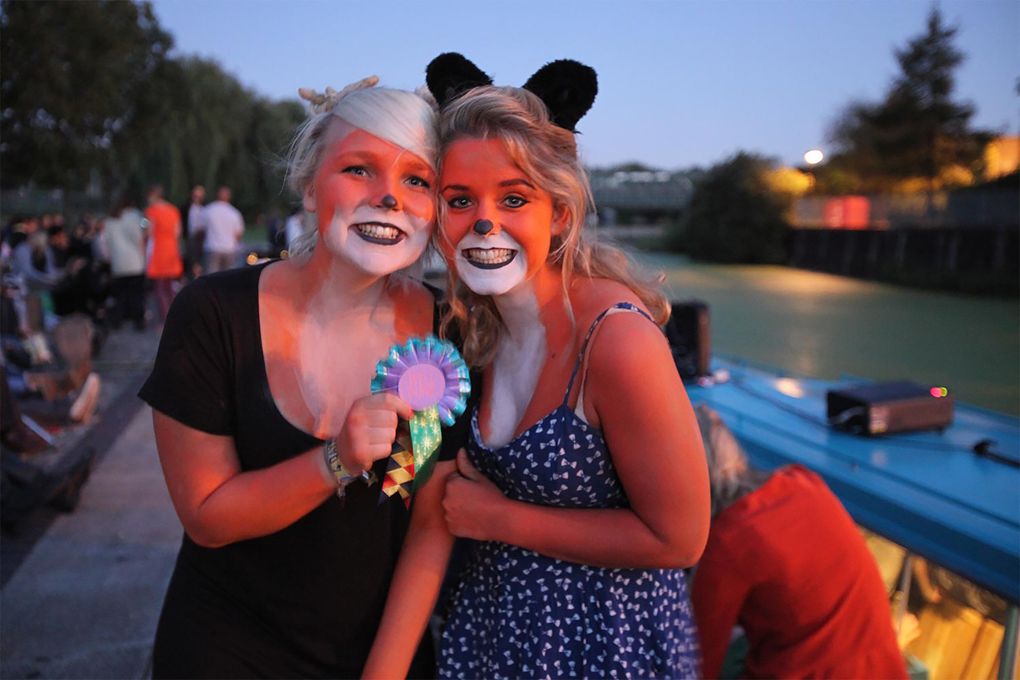 Two women with body and face paint to look like foxes, holding a blue ribbon badge next to a blue barge canal boat at dusk.
