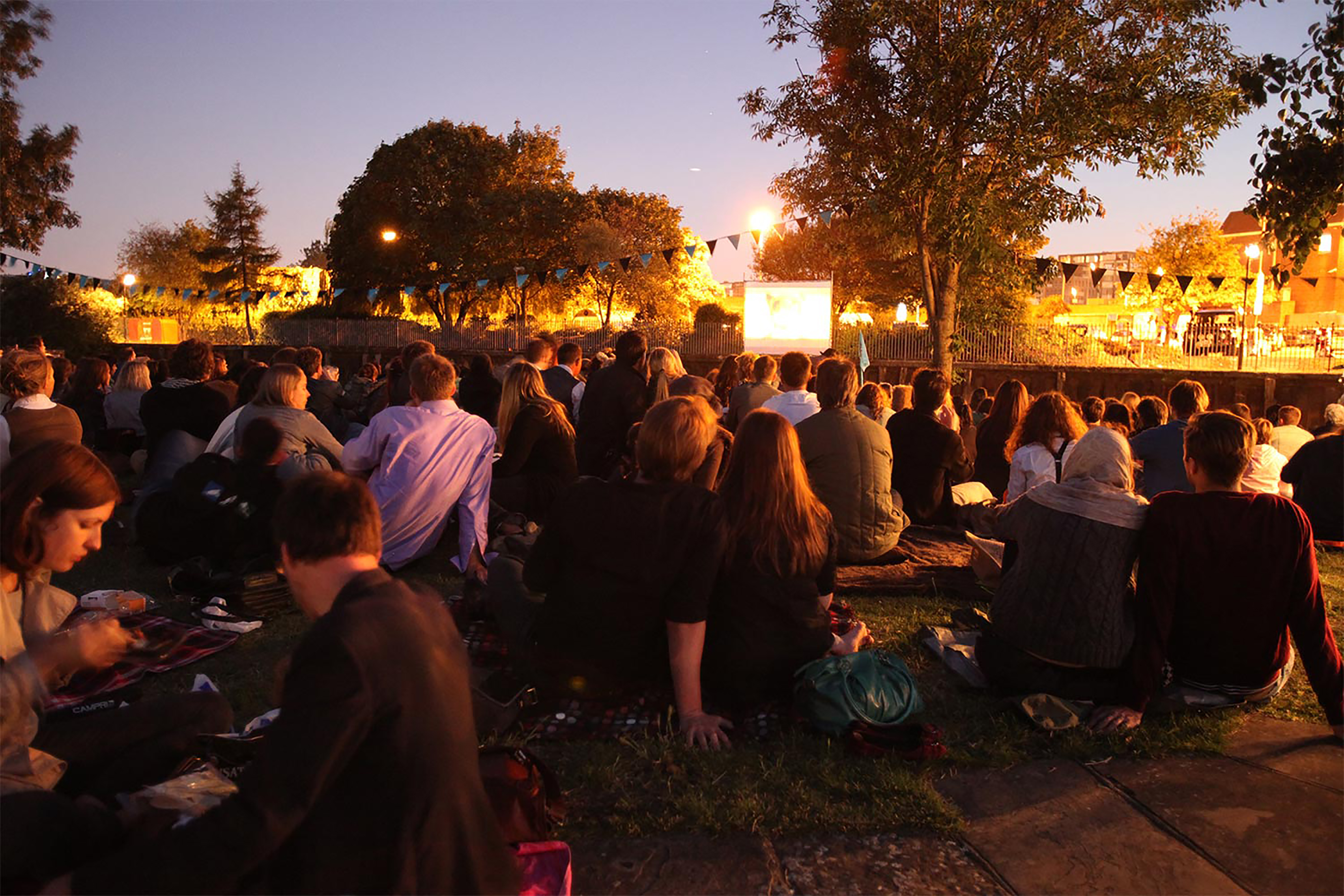 A large group of people at night all sitting on the ground facing towards a projection screen showing a movie at night, with hanging lights all surrounding them.