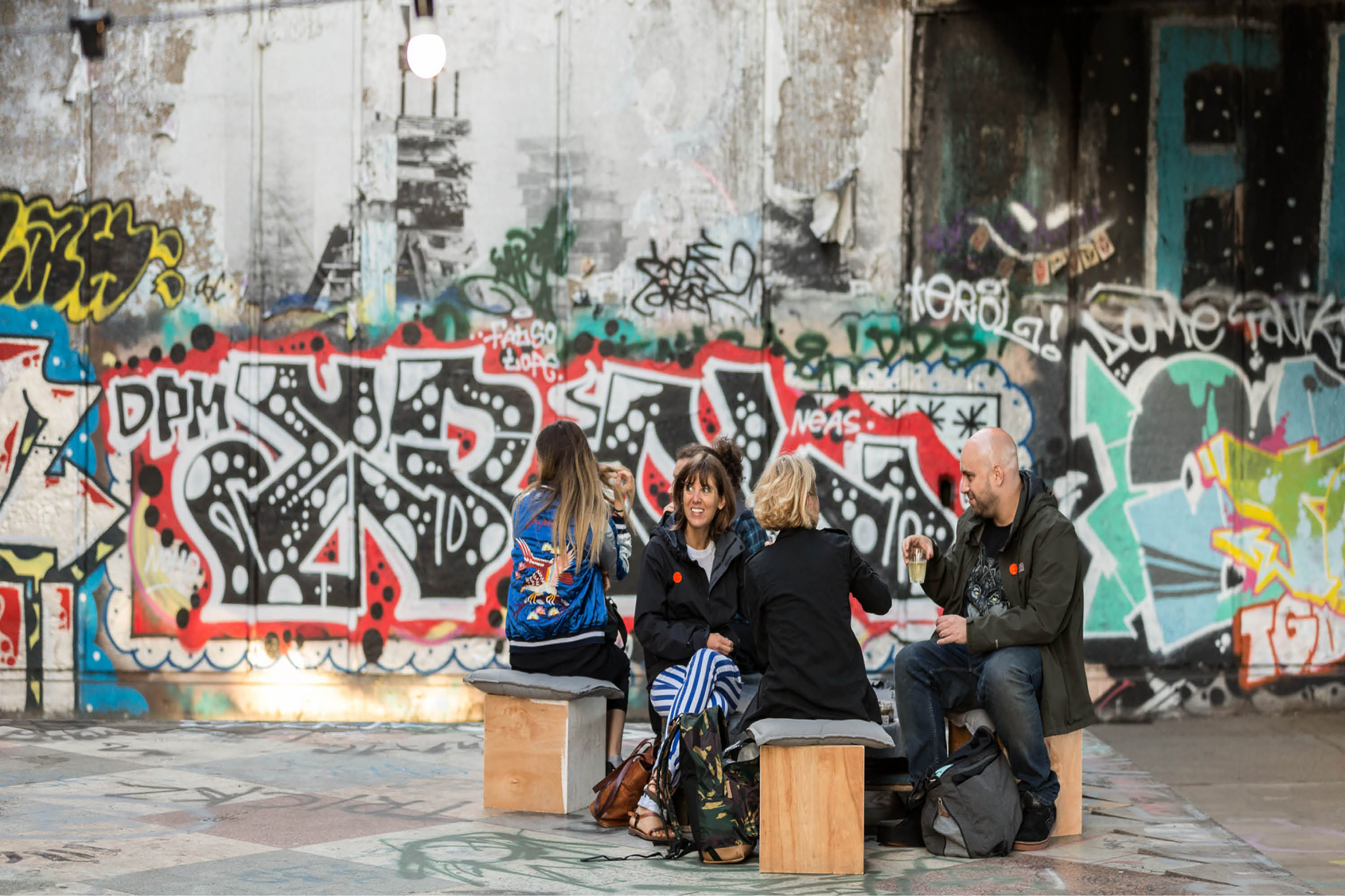 A group of people sitting on wooden blocks in a circle in front a large grey wall covered in graffiti.