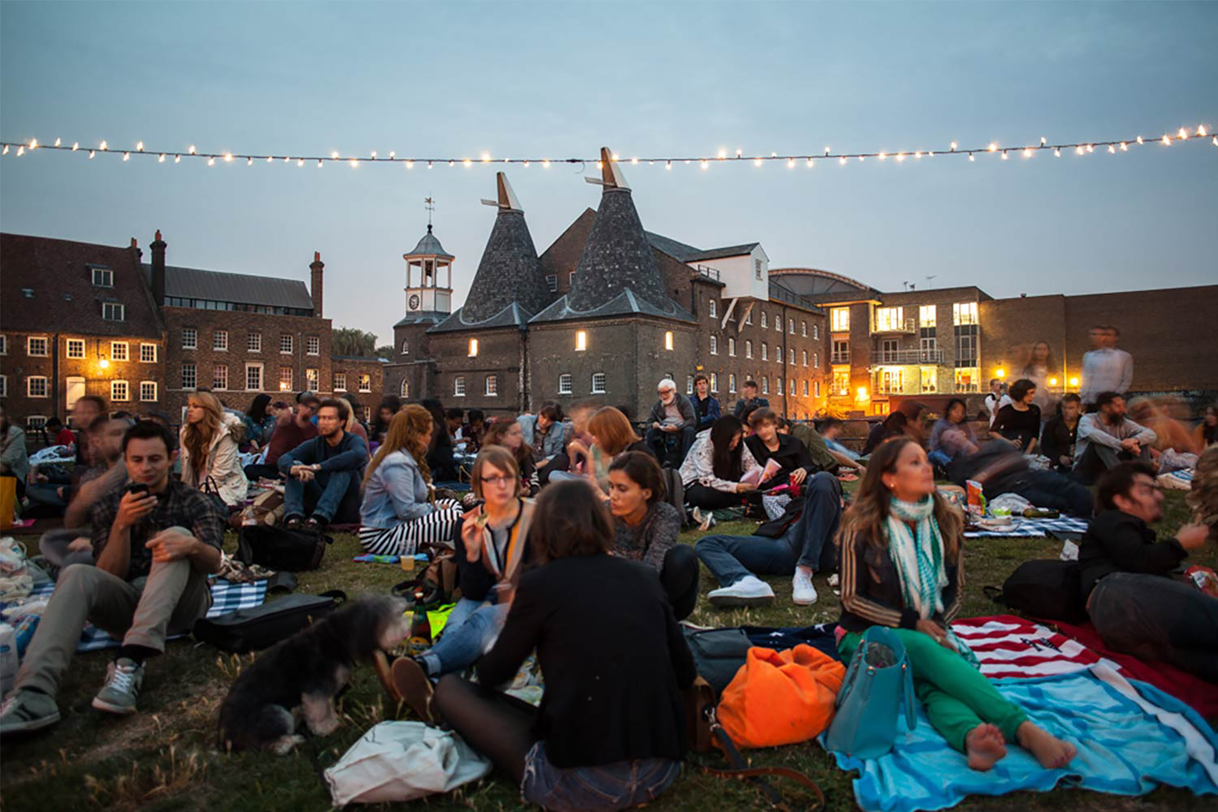 A large group of people at dusk all sitting on the ground facing towards a projection screen, with hanging lights all surrounding them.